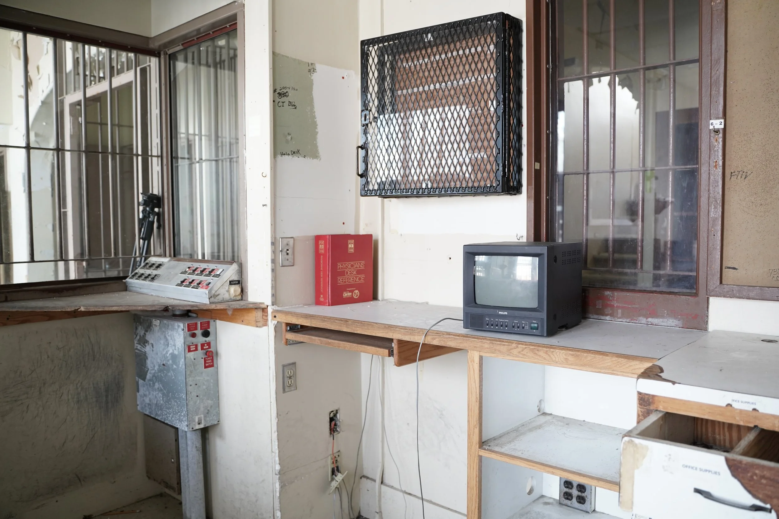 A construction or renovation site interior with a partial wall, an old CRT television, a red reference book titled 'Physicians Desk Reference', a black cable lock-covered window, an open electrical panel, and unfinished wooden shelves.