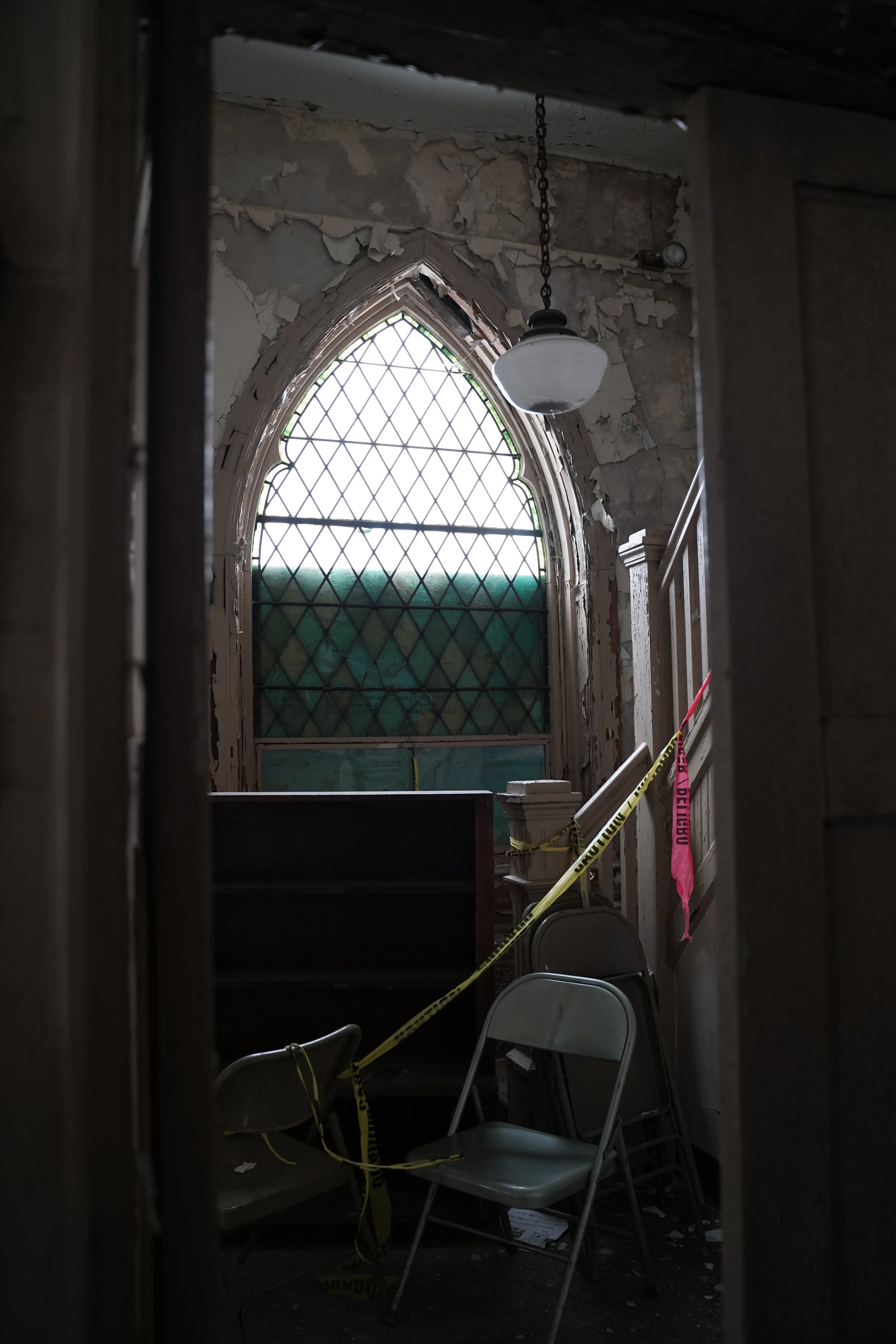 Image of an interior space with an arched stained-glass window, peeling paint on the walls, a hanging lamp, caution tape, and chairs, suggesting a space under renovation or disrepair.