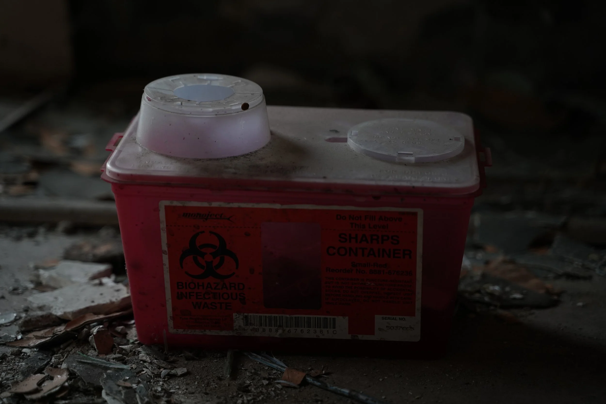 A red biohazard container with a biohazard symbol and warning labels, placed on a dusty, debris-covered floor in a dark environment.