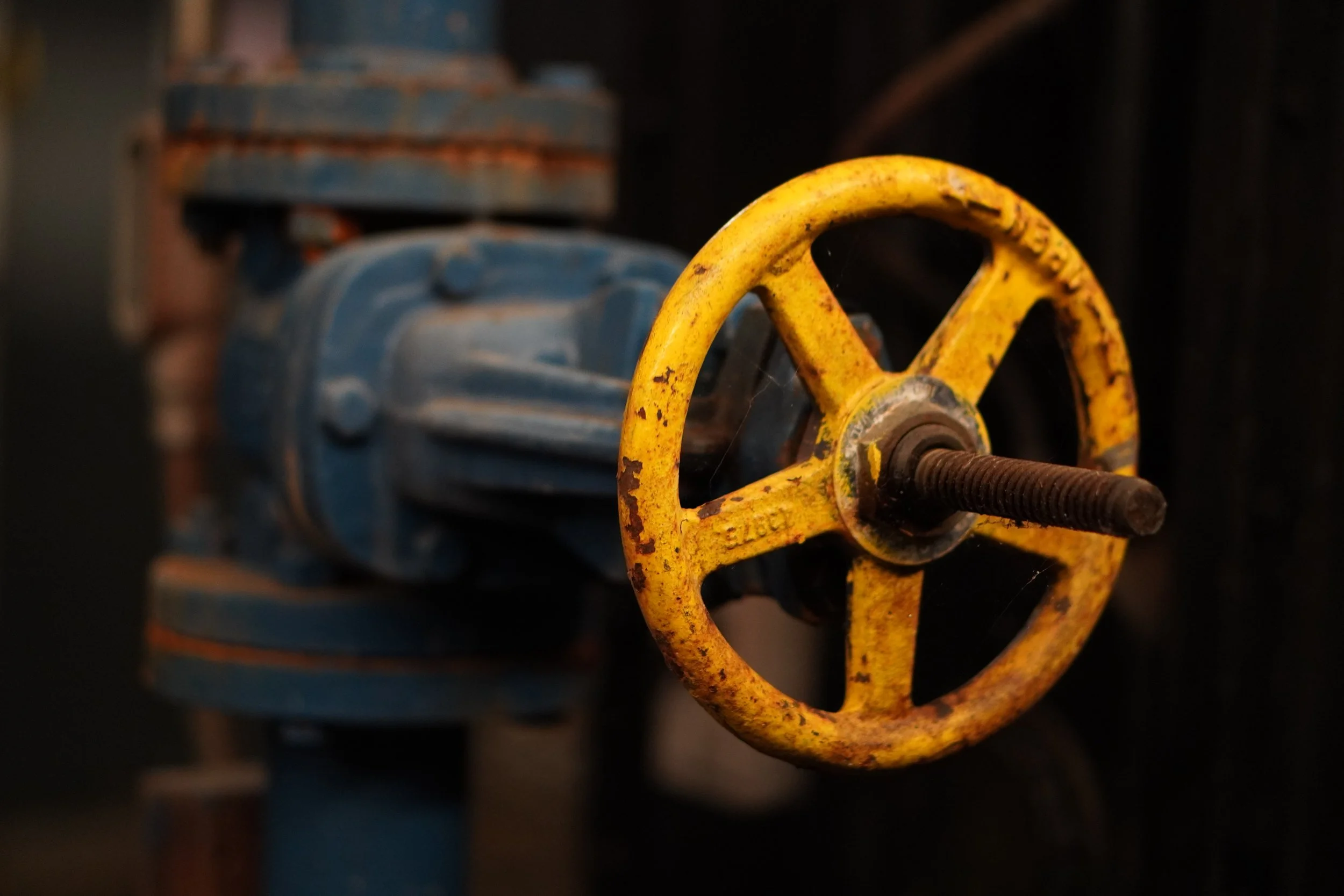 Close-up view of a yellow industrial valve handle with rust, in front of a blue valve. The background features other equipment and is out of focus.