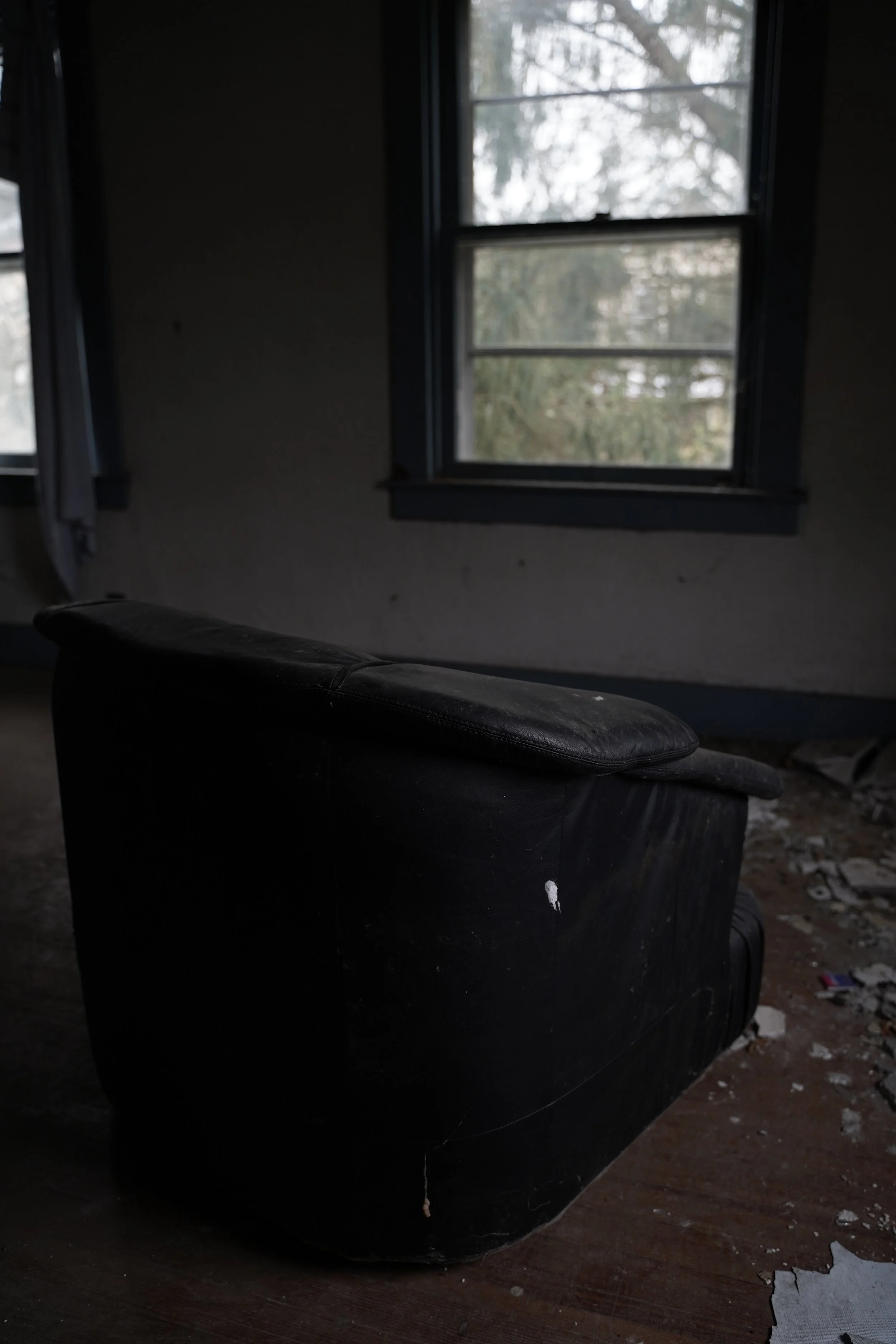 A dark, worn armchair in a dimly lit room with dirt and debris on the floor, and a window showing an overcast sky and trees outside.