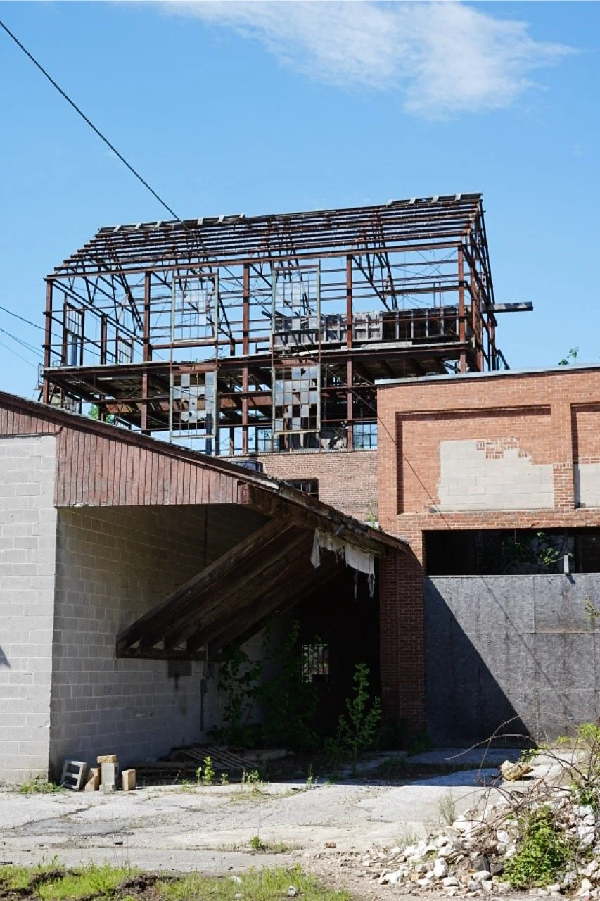 An abandoned building under construction or renovation with exposed steel framing on the top floor, brick walls, and overgrown weeds, with a partly collapsed roof and debris on the ground.