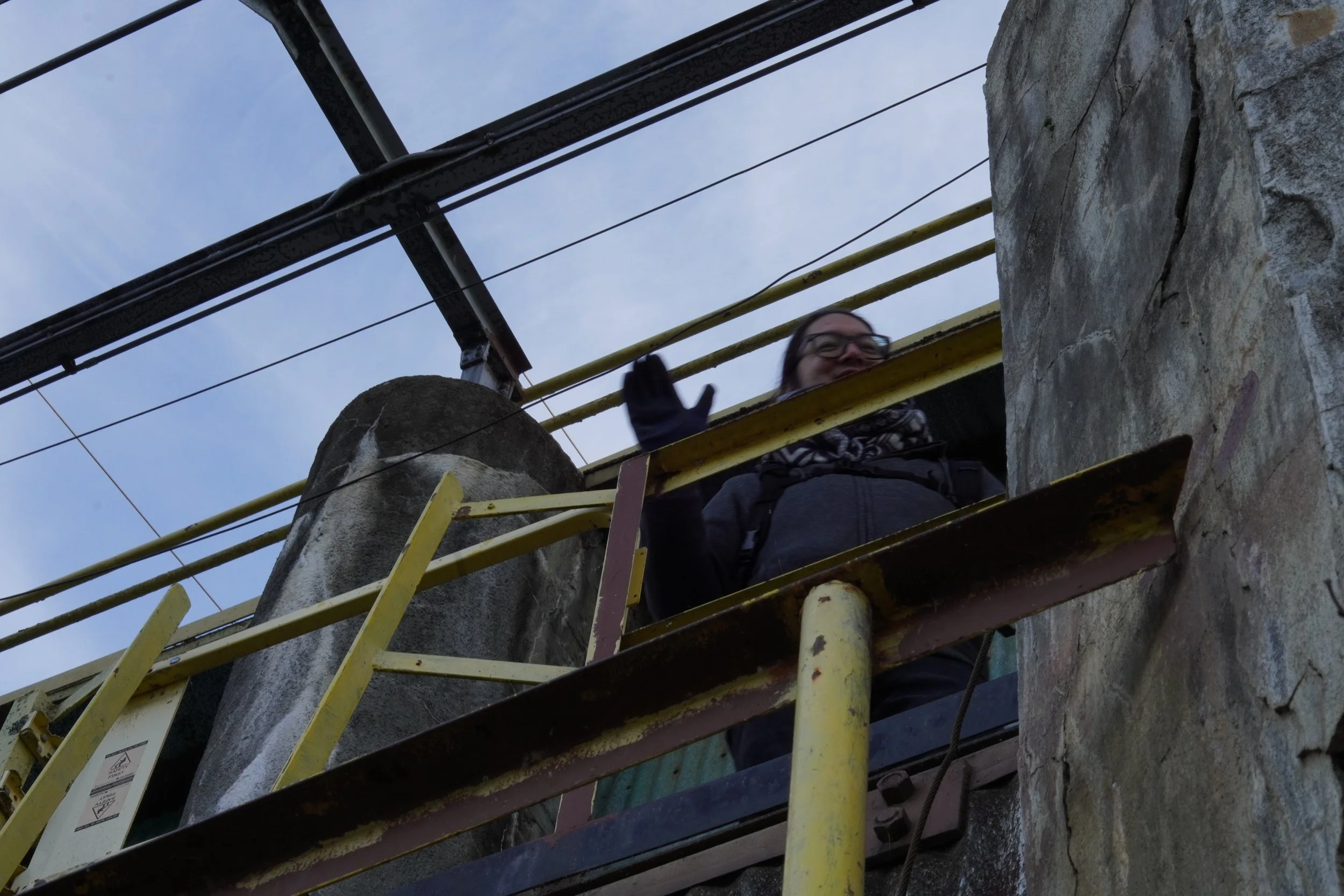 Person with glasses waving from a bridge or platform near a stone wall, with metal railings and electrical wires overhead.