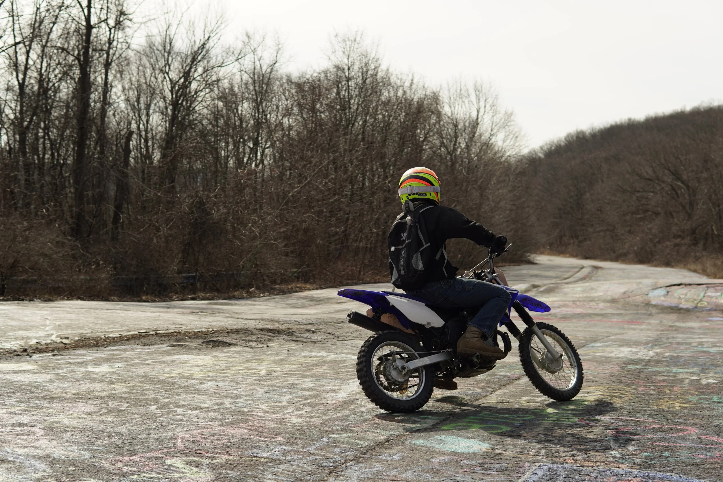 Person wearing a multicolored helmet riding a dirt bike on a paved surface with leafless trees in the background.