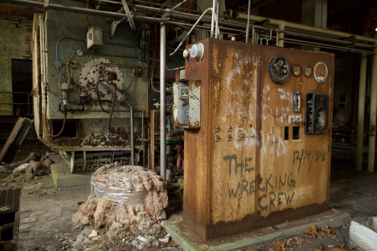 An abandoned, rusted industrial control panel with the words "THE WRECKING CREW" spray-painted on it, surrounded by debris and rusted machinery.