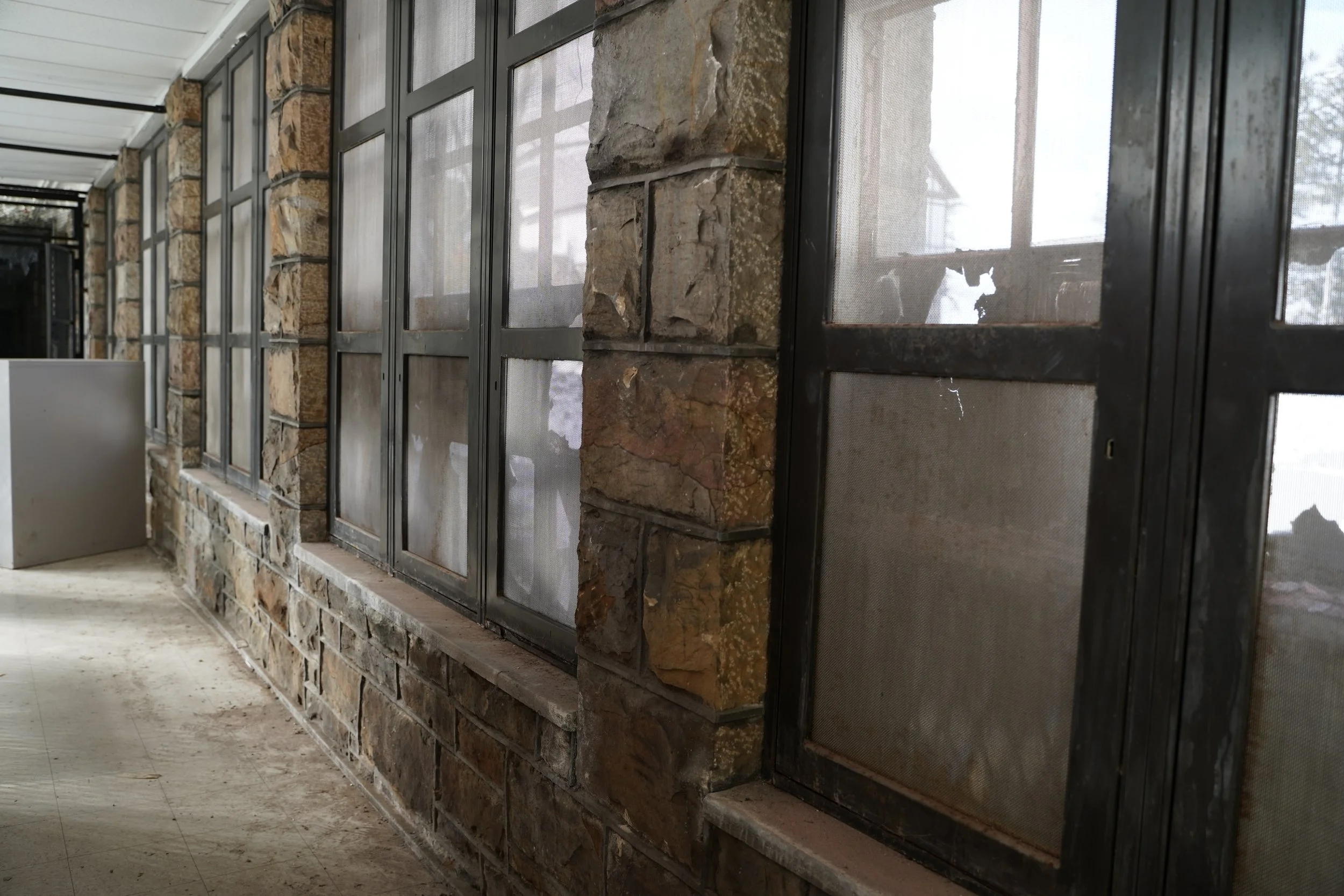 A stone wall with multiple black metal-framed glass doors or windows, some with screens, is shown. There is a concrete floor and a white ceiling.