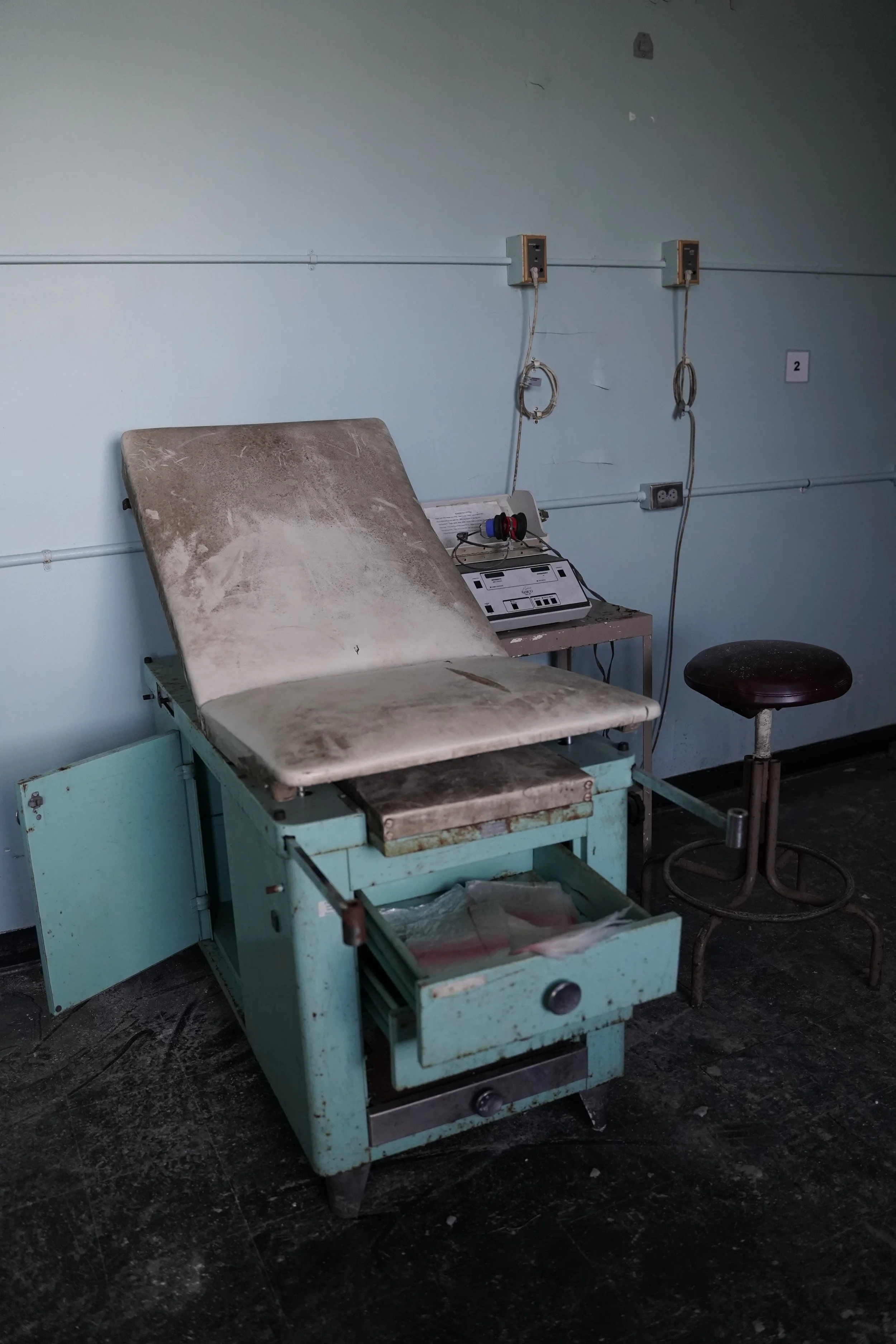An empty hospital or medical examination room with an old, dirty medical examination table, a stool, and some medical equipment on a counter, with peeling paint and worn-out walls.