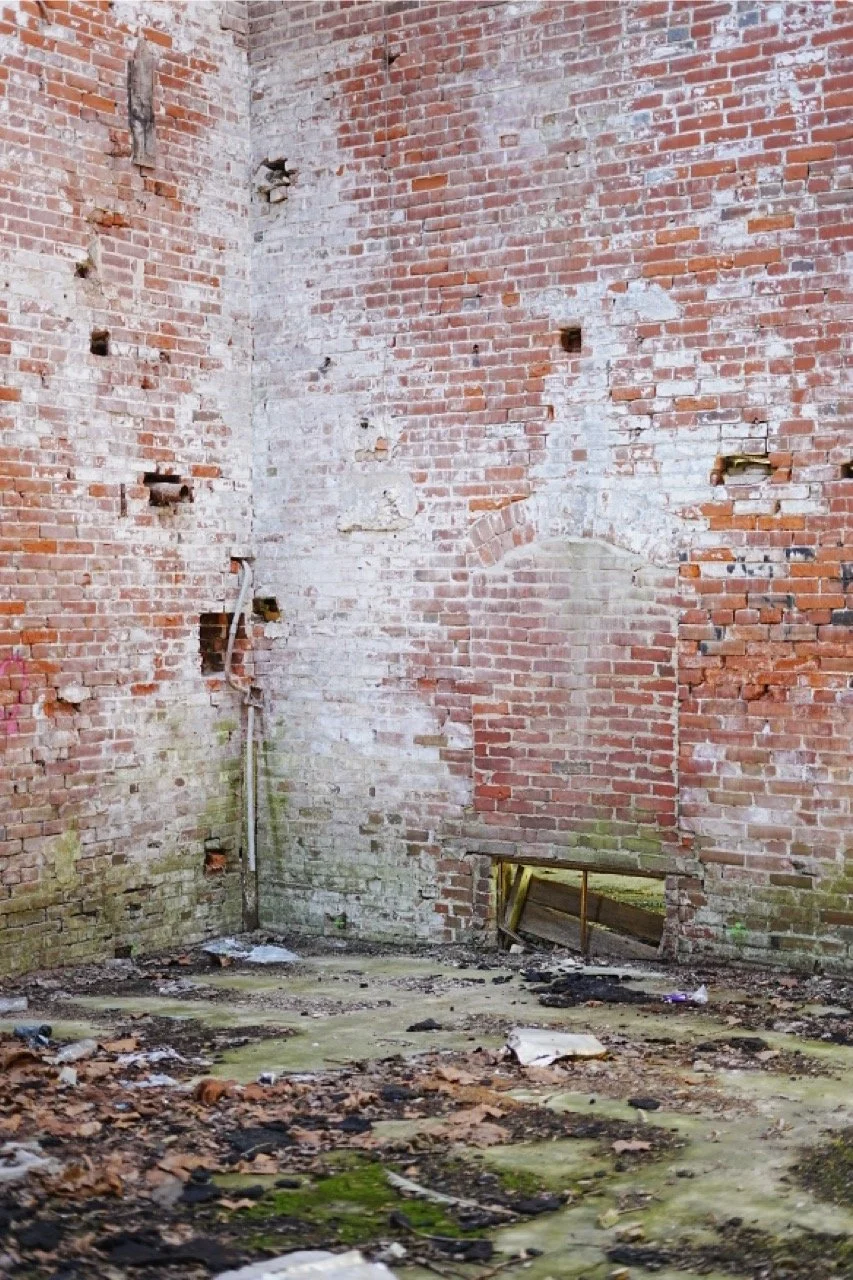 An empty, abandoned brick room with damaged walls and debris on the floor. There is a small rectangular opening near the bottom of the wall, possibly a vent or window, with wooden supports underneath.
