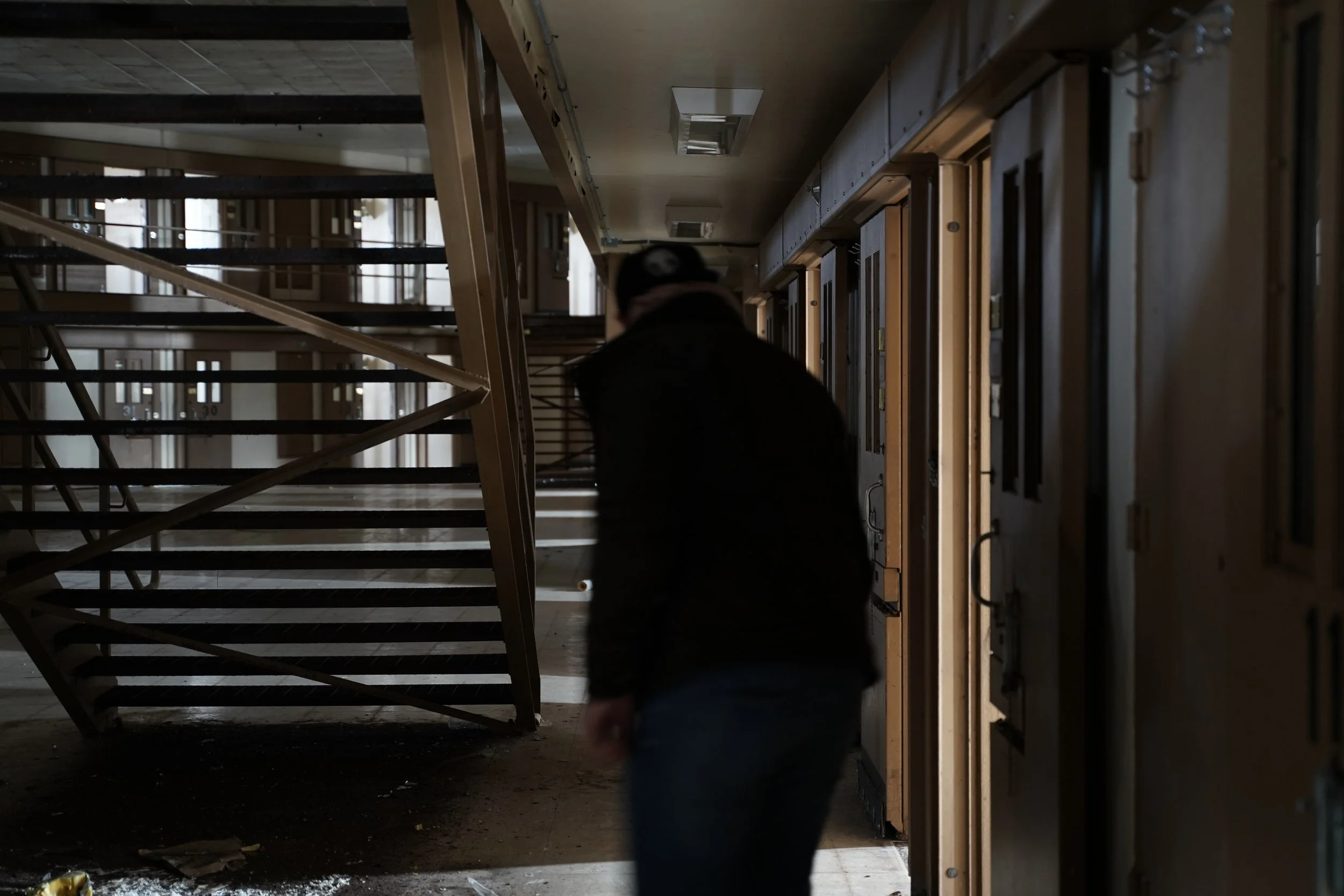 A person walking through a dark, abandoned building with broken floors, exposed structures, and boarded-up windows. The scene is dimly lit, creating a moody and eerie atmosphere.