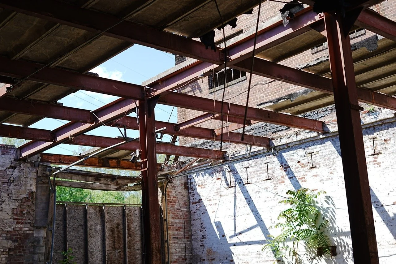 The interior of a building under construction with exposed brick walls, steel beams, and some plants growing inside.
