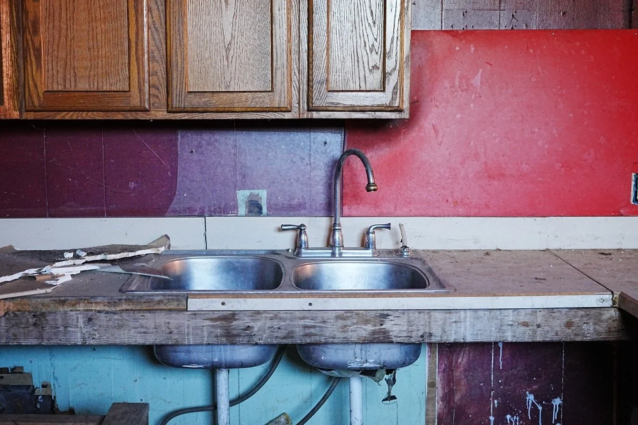 Dirty kitchen sink with a broken pipe and peeling paint on the countertop and walls, upper wooden cabinets, and partially exposed plumbing.