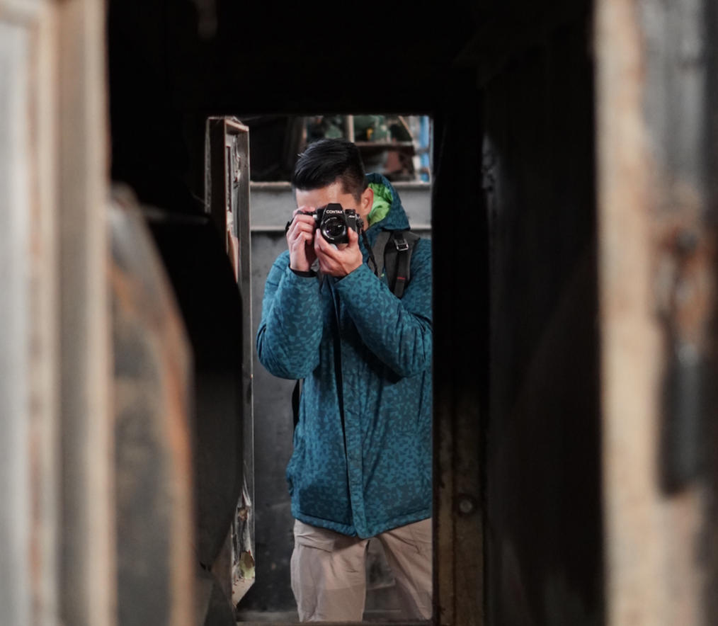Person in blue jacket with backpack taking a photograph through a narrow, dark passageway.