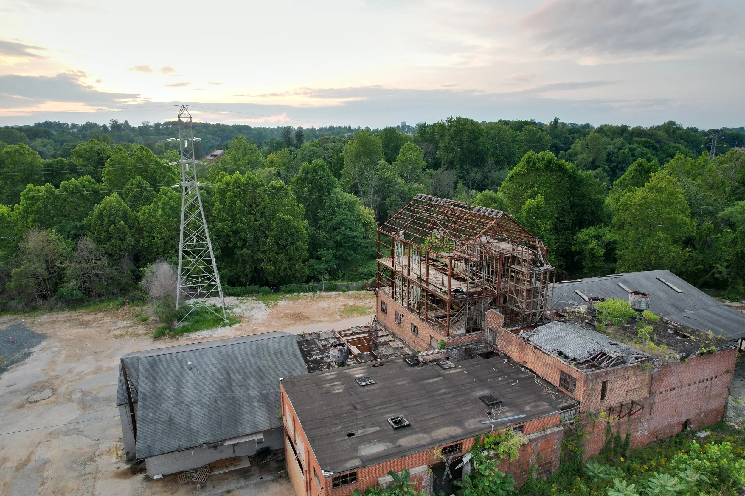 Aerial view of a construction site with a partially built brick building, surrounded by greenery and trees. An electrical tower and power lines are visible, with a cloudy sky overhead.