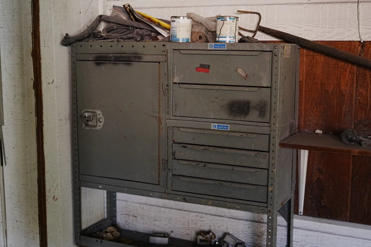 A gray metal tool cabinet with multiple drawers, some with black stains, standing against a white and brown wooden wall. The top of the cabinet holds three paint cans, a cloth, and various tools and materials. A small wooden shelf to the right has a 