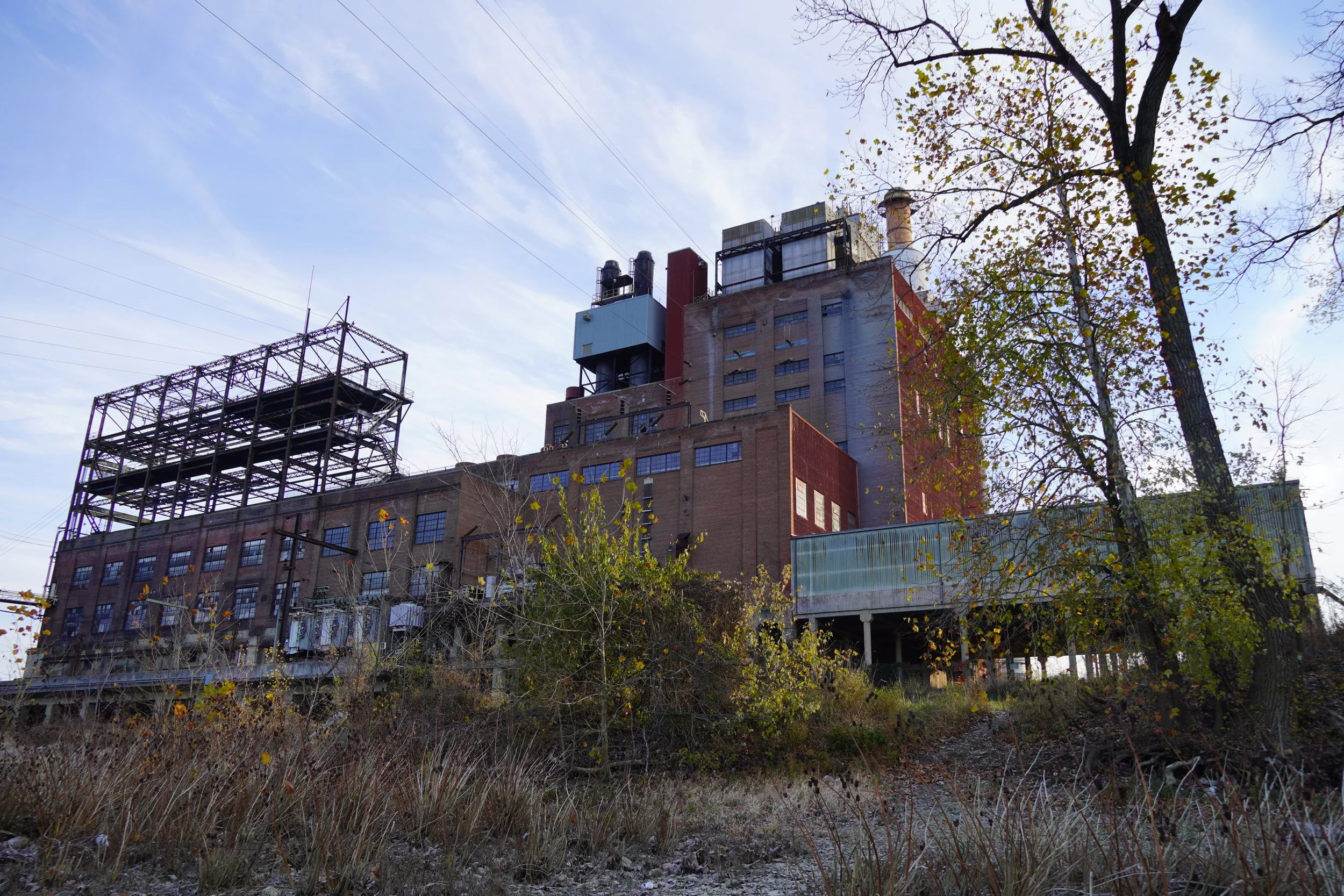 An industrial building with various structures on its rooftop, including chimneys, HVAC units, and metal frameworks, is visible behind trees with sparse autumn leaves. Power lines run across the sky above the building, and there is a mix of natural a