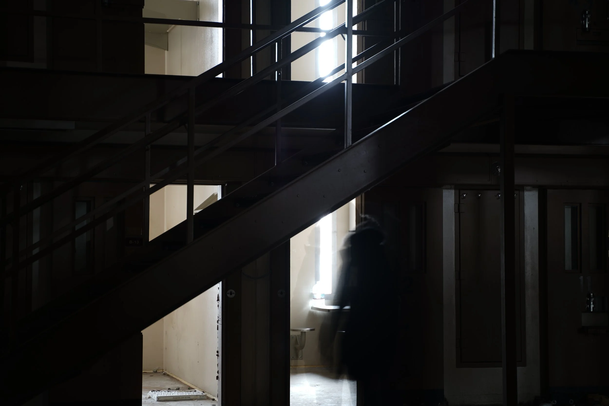 Dark interior of a building under construction with metal stairs and partially finished walls, illuminated by natural light coming from an open door and windows.