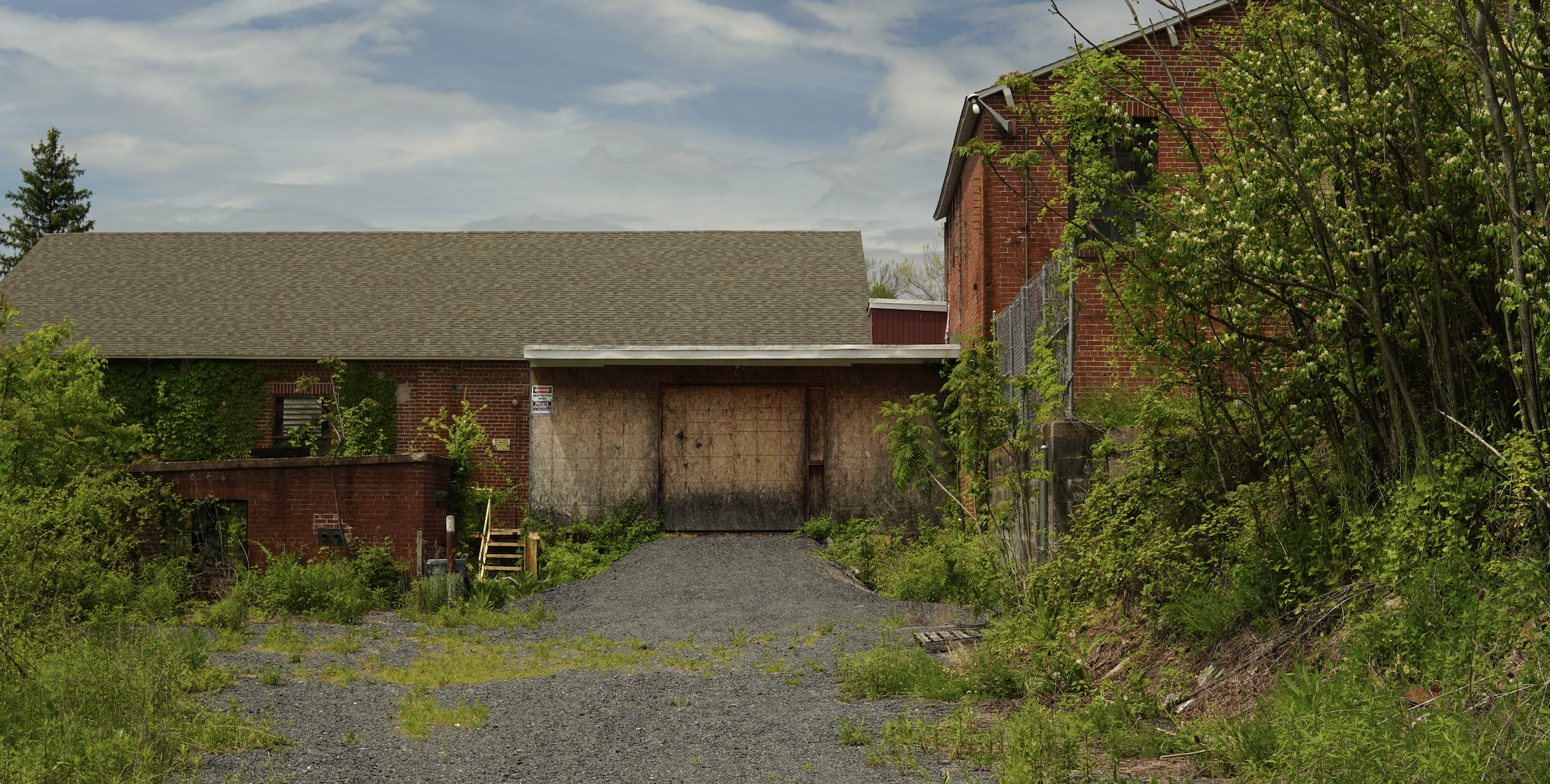 A derelict brick building with boarded-up windows, overgrown vegetation, and an uneven gravel driveway in front, under a cloudy sky.