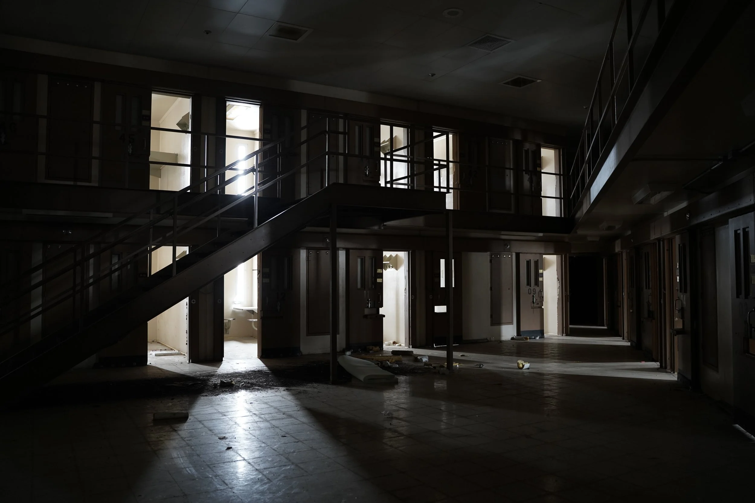 Dimly lit, abandoned interior space with broken tiles on the floor, open elevator doors, a staircase leading upward, and debris scattered around.