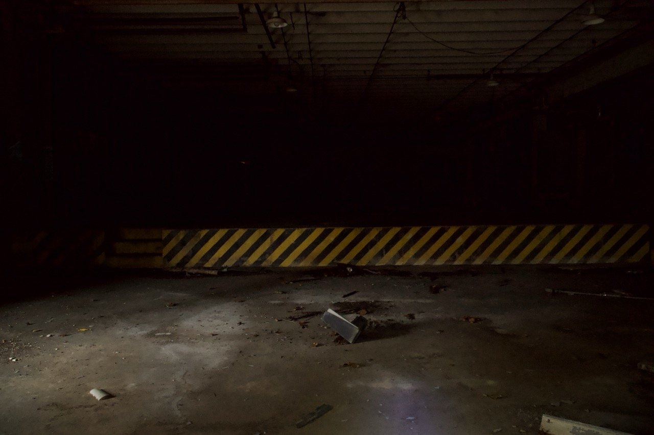 Dark, abandoned parking garage with a yellow and black striped barrier and scattered debris on the floor.