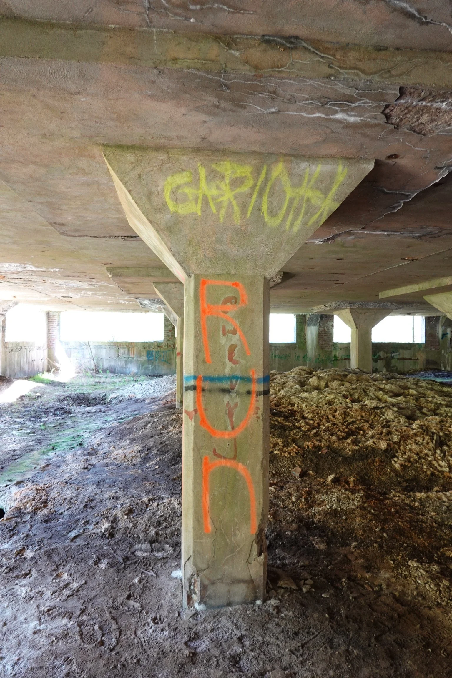 Concrete column supports an elevated structure in an unfinished basement or parking garage, covered with graffiti tags in yellow and orange paint. The floor is uneven dirt and rocks, with sunlight coming in from openings at the background.