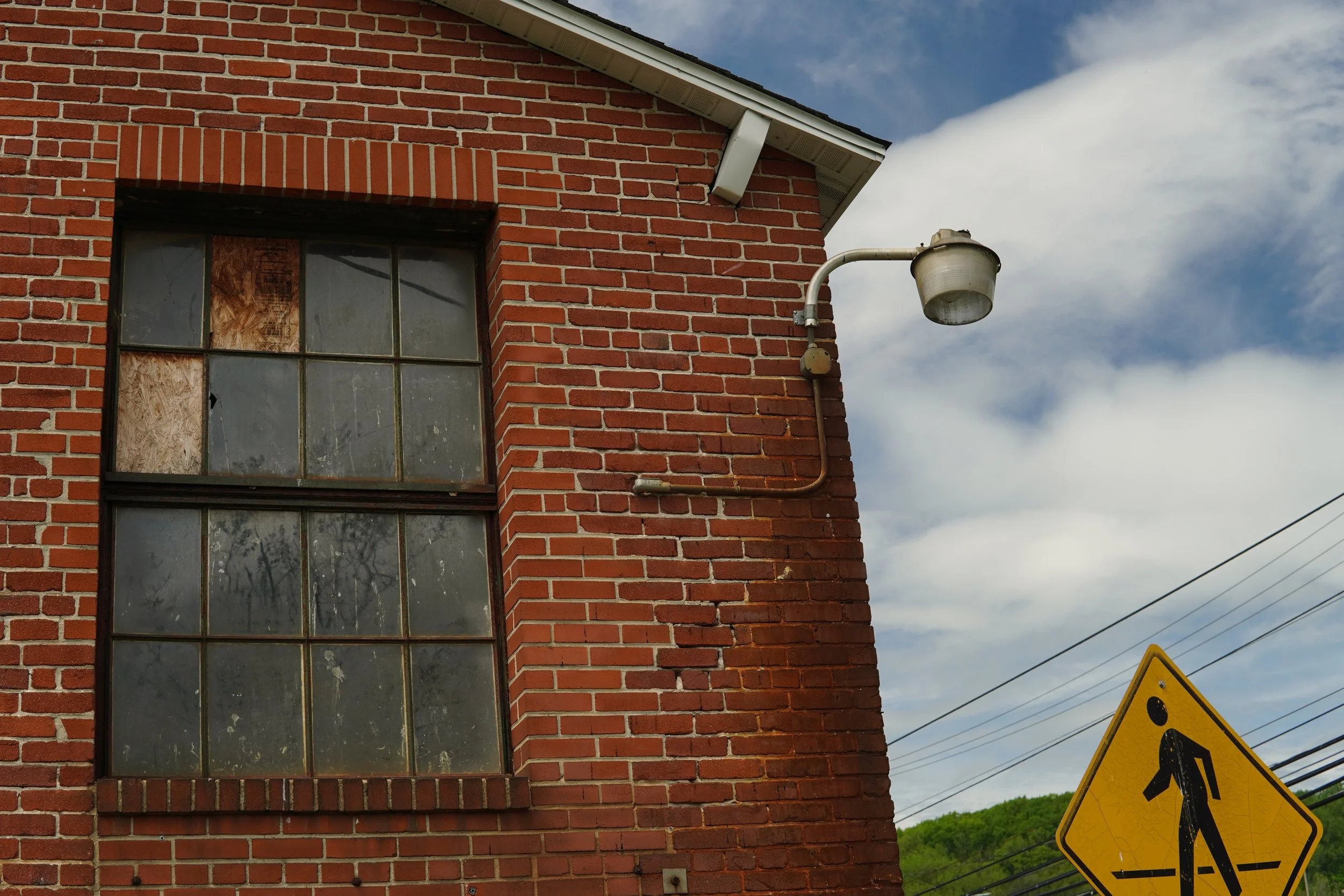 Red brick building with a large window and an outdoor light fixture attached to the wall. A caution sign is partially visible in the bottom right corner, and the sky with clouds is in the background.