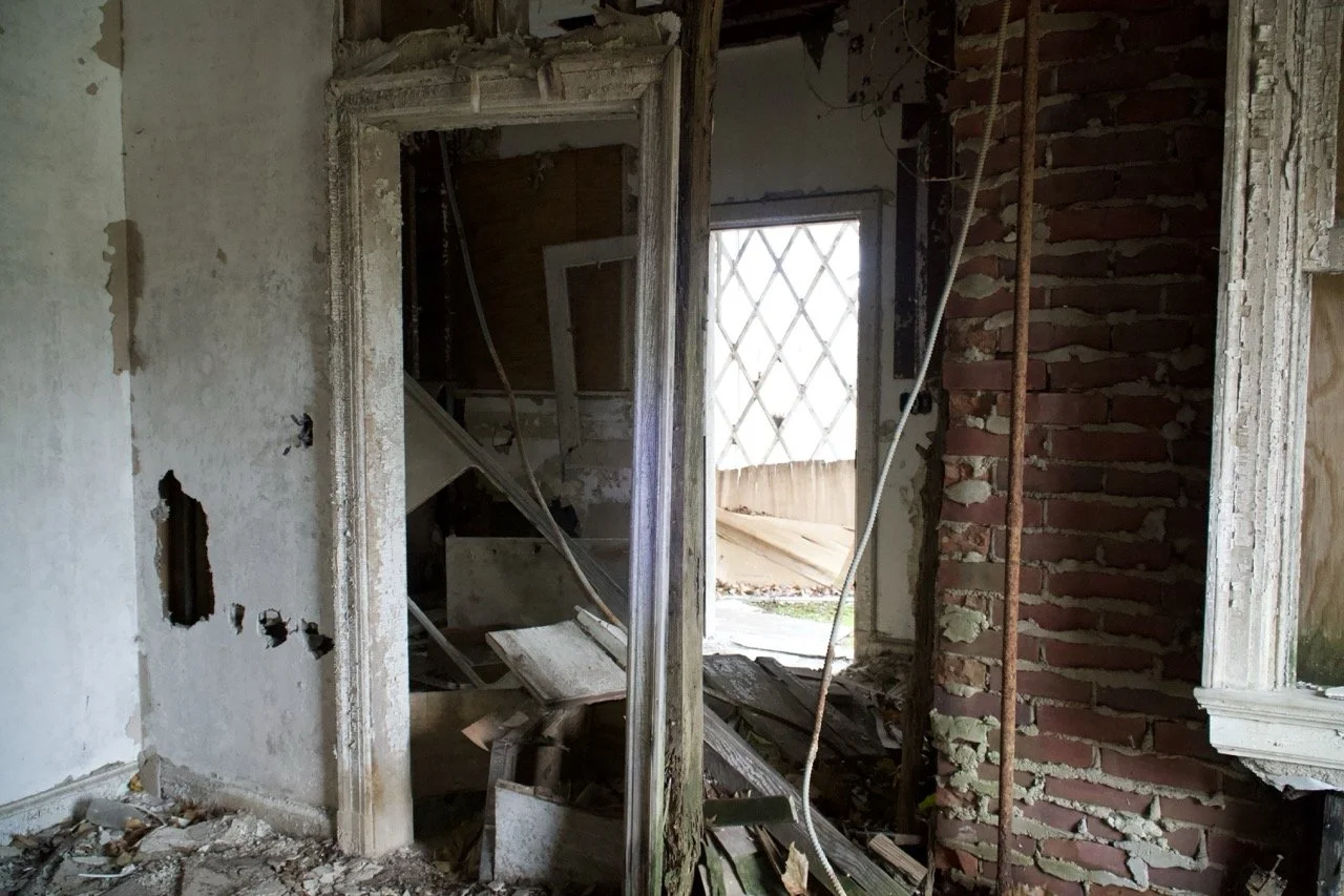 Interior of a dilapidated building with damaged walls, exposed bricks, and debris on the floor; a door frame without a door and broken window provide a view of the outside.