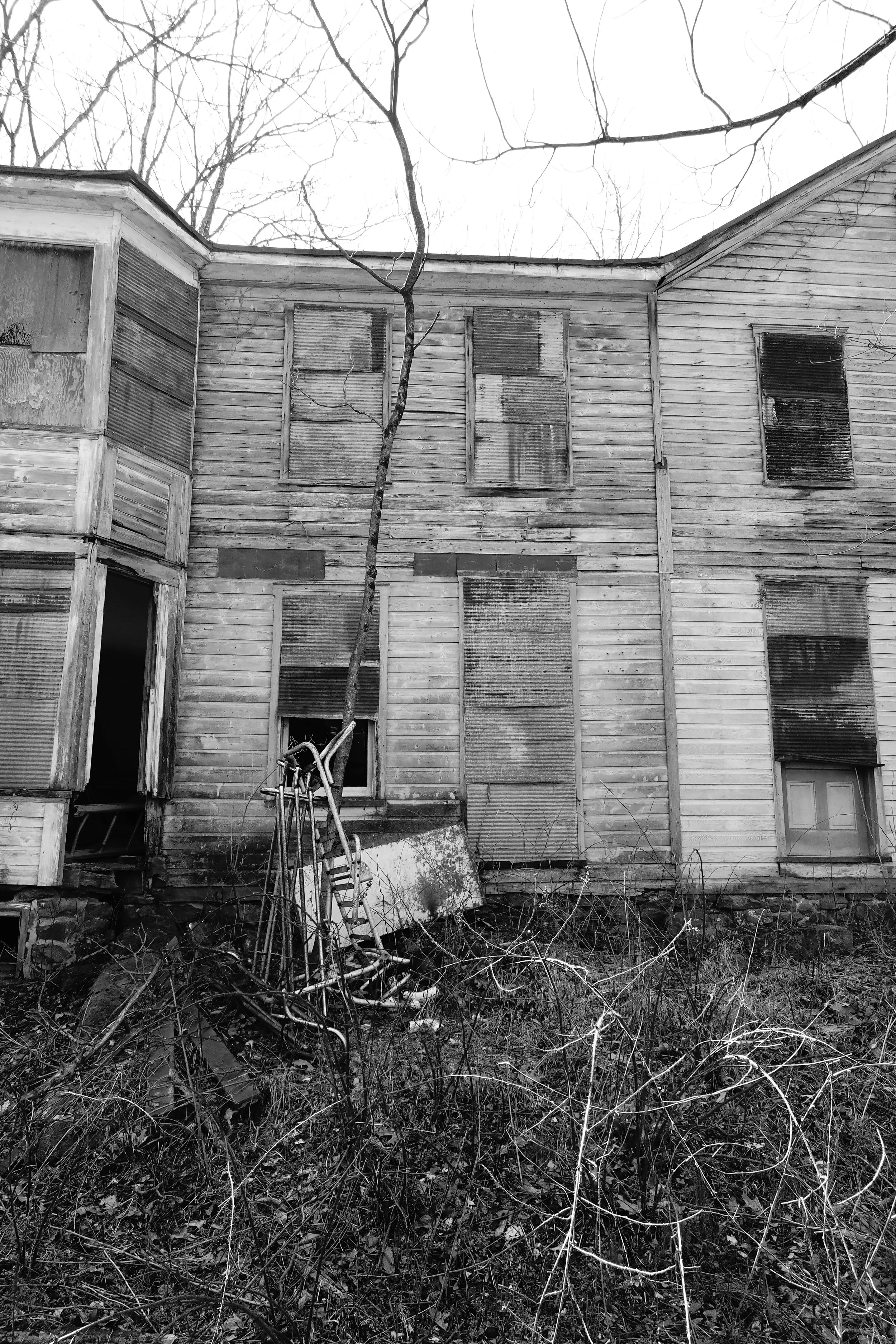 An old wooden house with boarded-up windows and missing glass, surrounded by overgrown weeds and leafless trees in a desolate setting, captured in black and white.