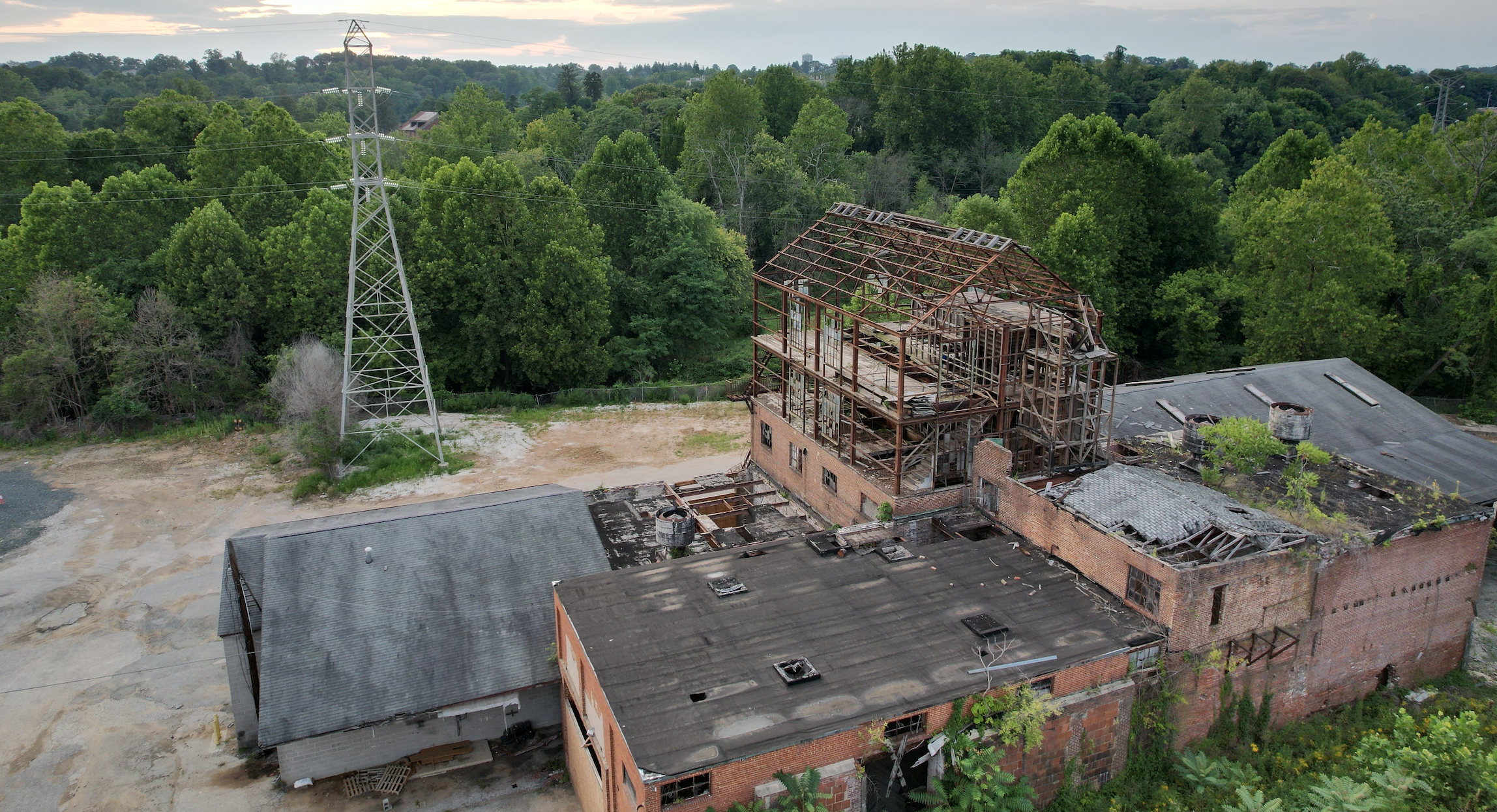 An abandoned brick building with a partially collapsed roof surrounded by trees, with a large metal power pole and green forest in the background.