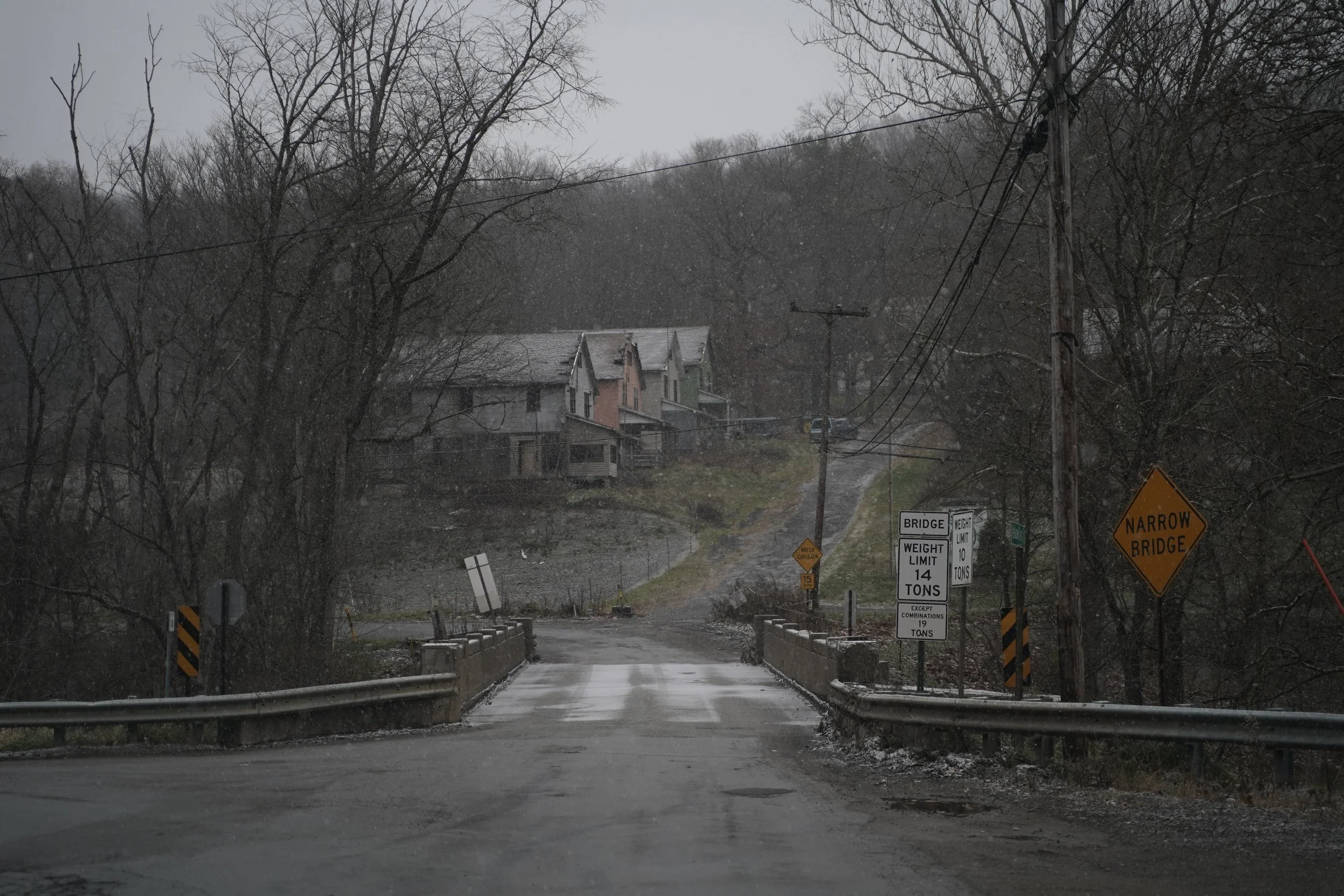A rural bridge over a small road on a rainy, overcast day with houses and trees in the background and multiple traffic and warning signs.