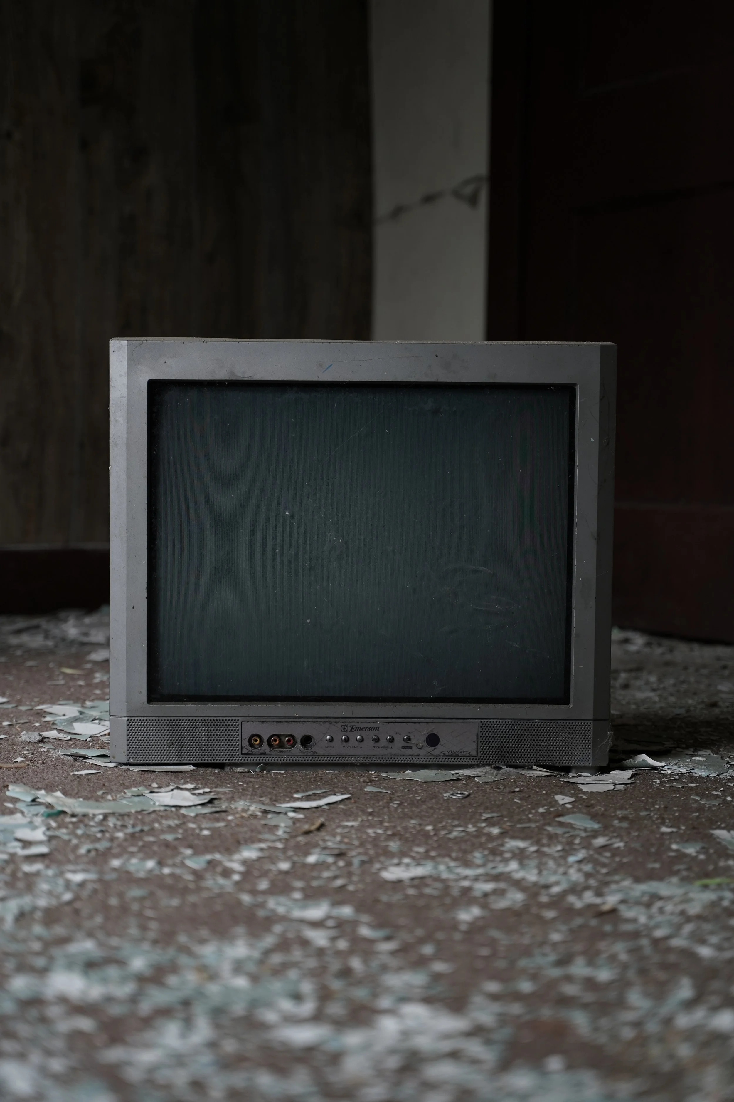Old, dusty computer monitor with a blank screen, sitting on a dirt floor surrounded by scattered debris and peeling paint from the wall behind it.