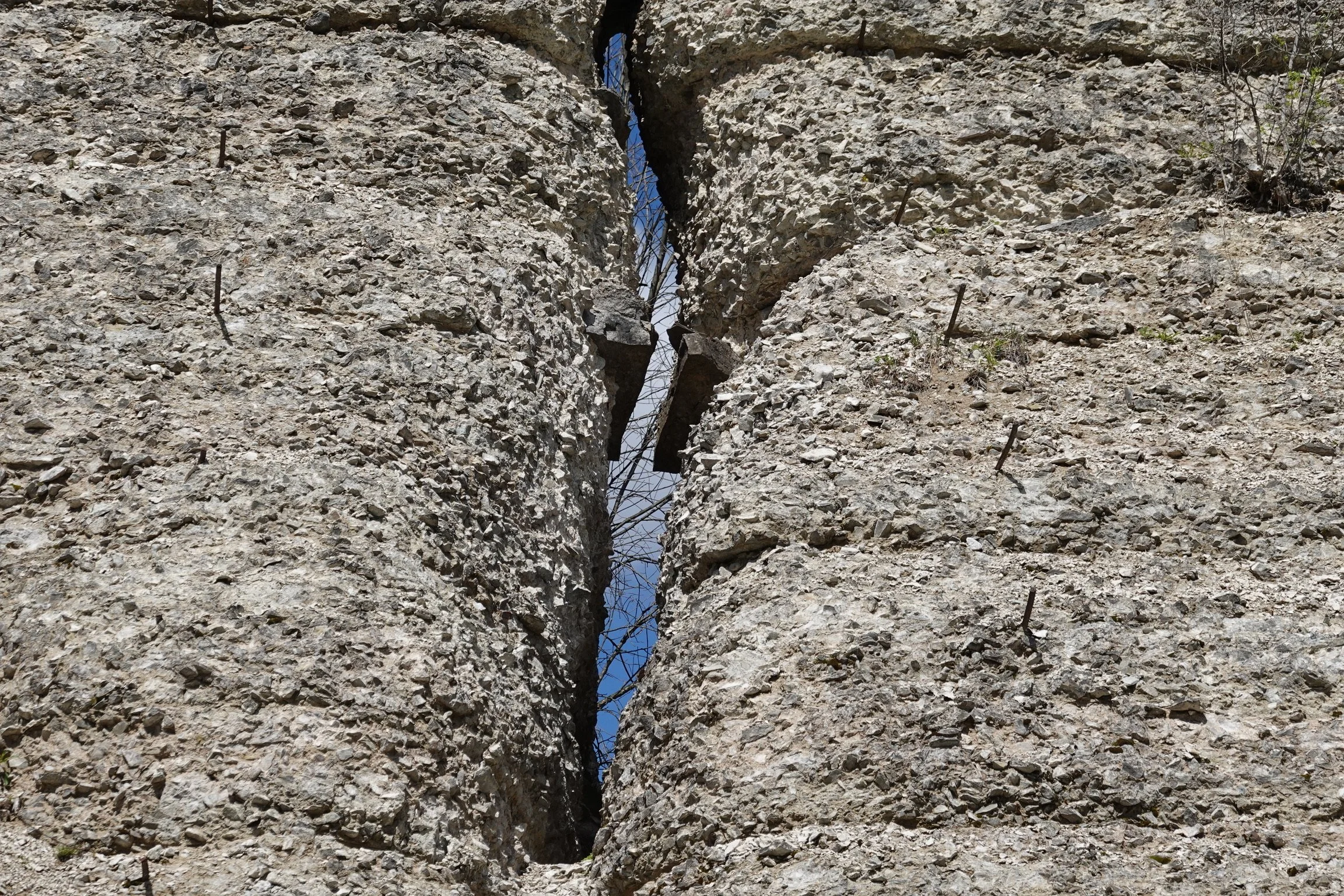 A close-up of a large concrete or stone wall with a deep crack running vertically down the center. There is some rusted bar reinforcement sticking out from the wall, and small patches of vegetation growing near the edges. The sky is visible through t