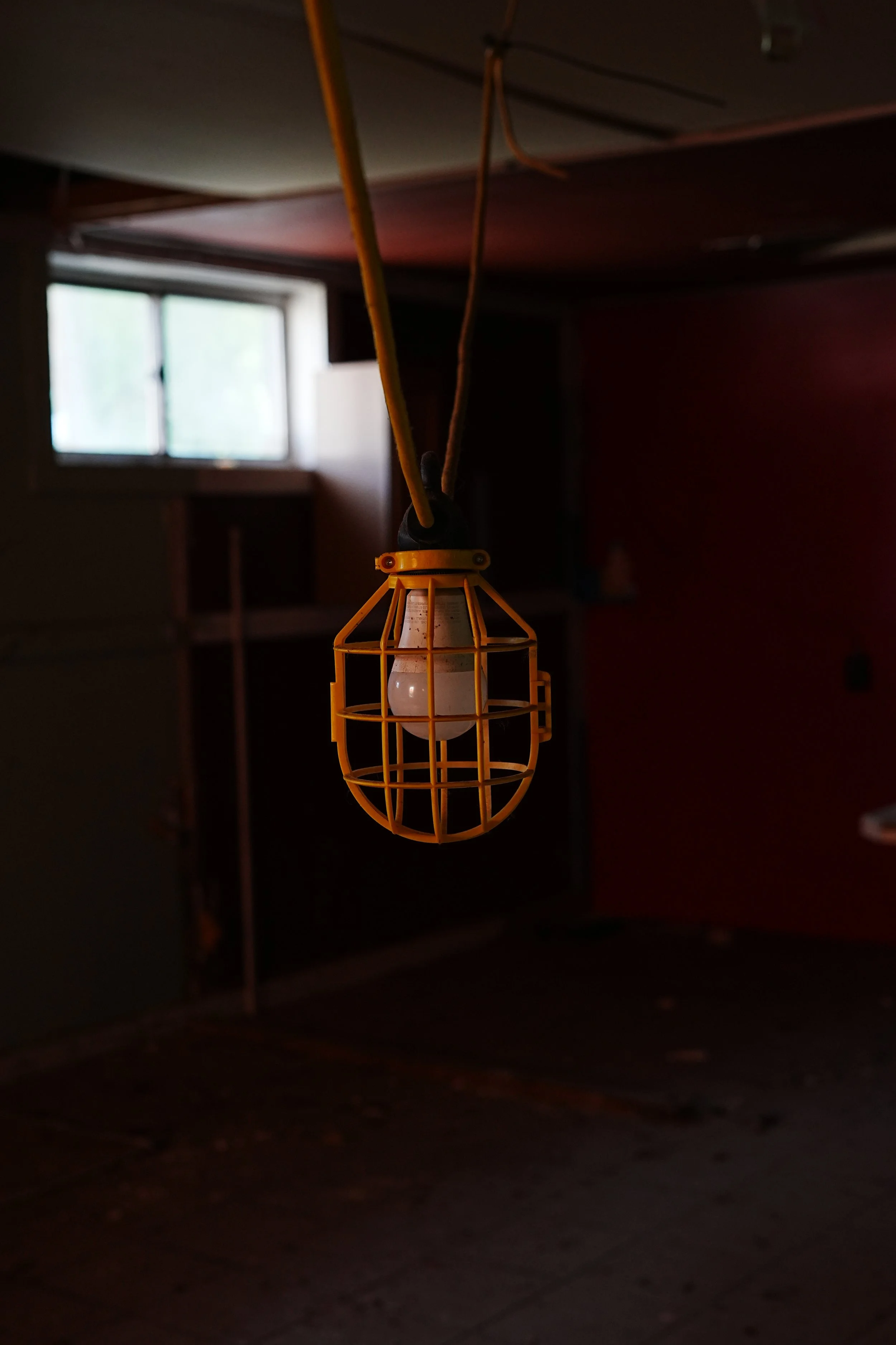A hanging yellow industrial-style cage light fixture with a white bulb inside, in a dimly lit room with a window in the background.