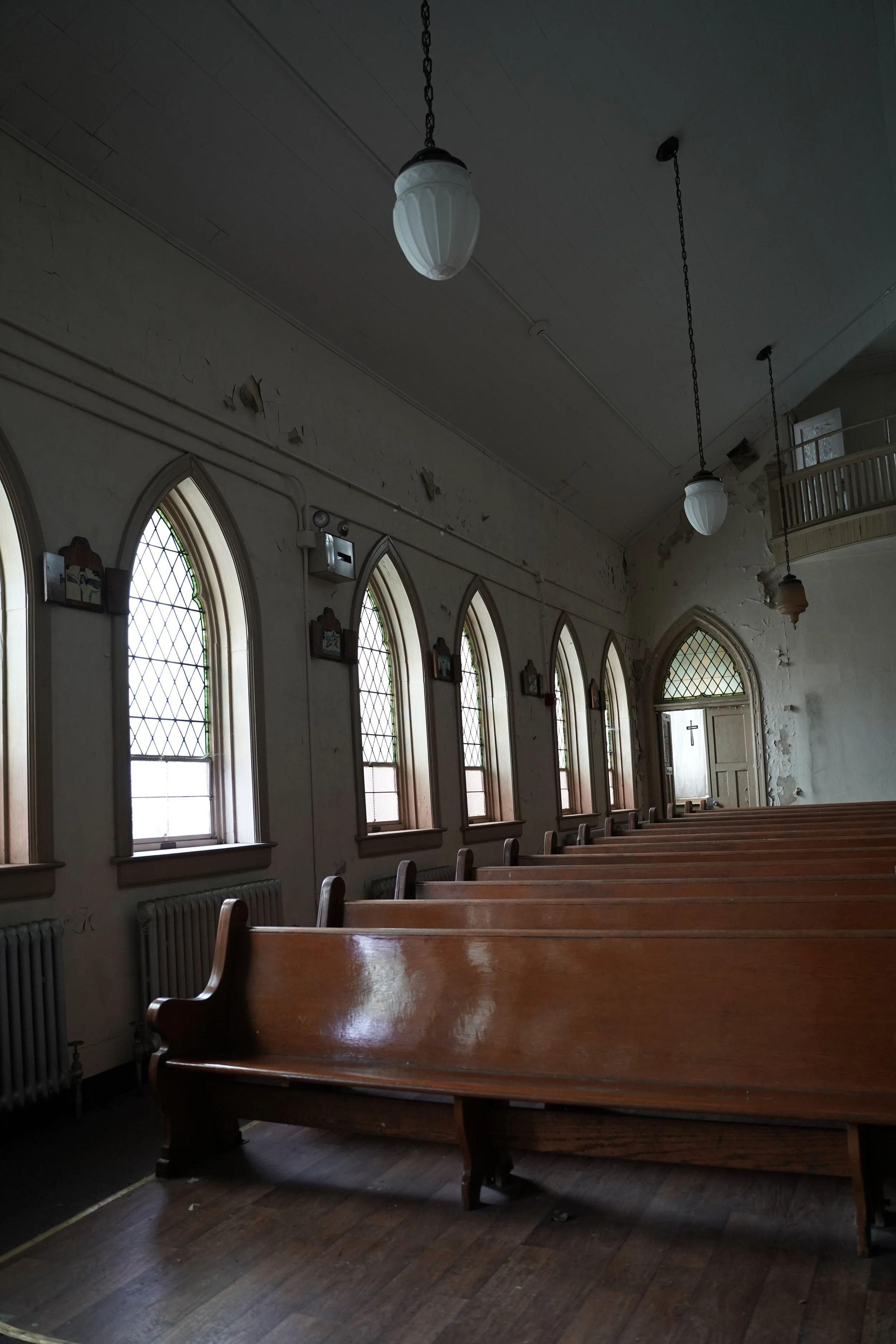 Interior of an old church with wooden pews, arched stained glass windows, and hanging light fixtures on the ceiling. The wall shows peeling paint or plaster.