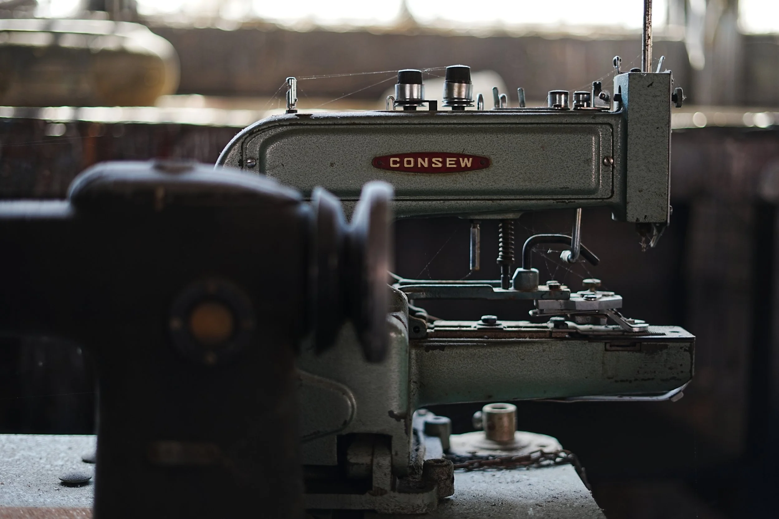 A vintage Consew sewing machine sitting on a surface, with a blurred background of what appears to be an industrial or workshop setting.