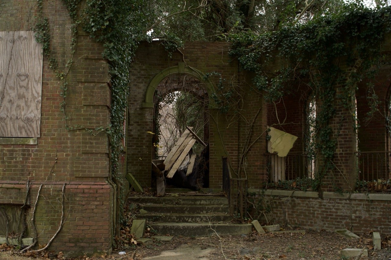 Abandoned brick building with boarded-up windows and overgrown ivy, front stairs cluttered with debris, broken wooden door leaning against doorway, surrounded by dead leaves and fallen branches.