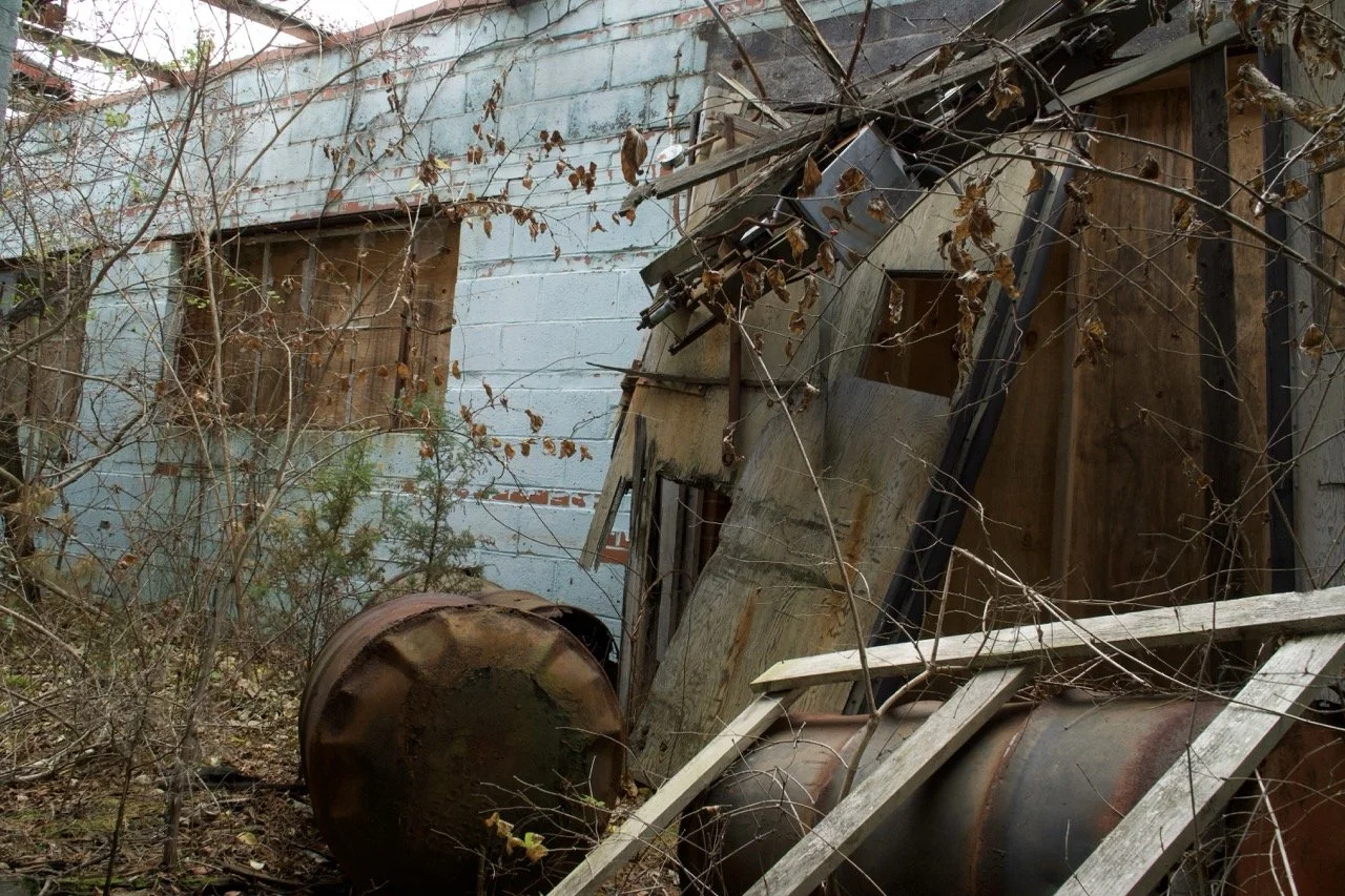 An abandoned, dilapidated building with boarded-up windows, overgrown with dead branches and foliage, rusted metal barrels, and broken wooden planks.