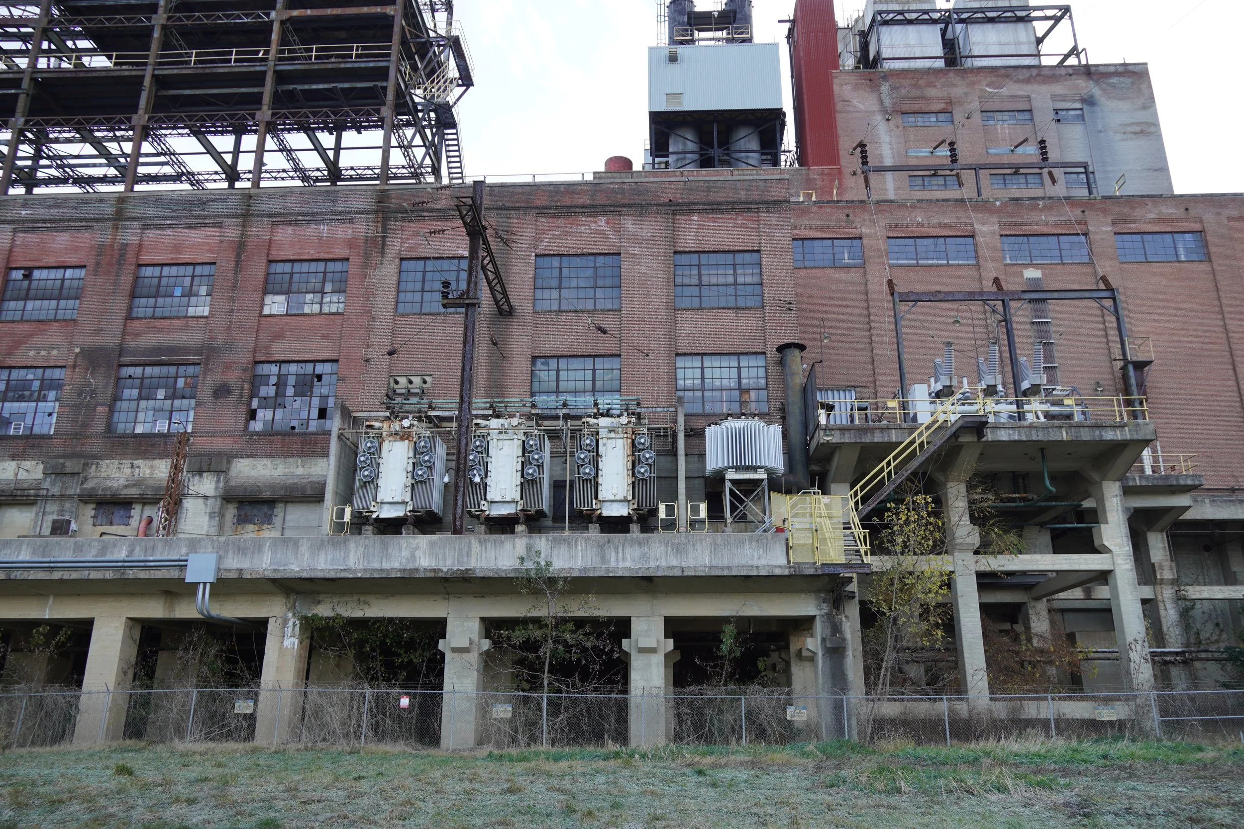 Back side of an old brick industrial building with large windows, electrical equipment, and metal framework, with a grassy area in the foreground.