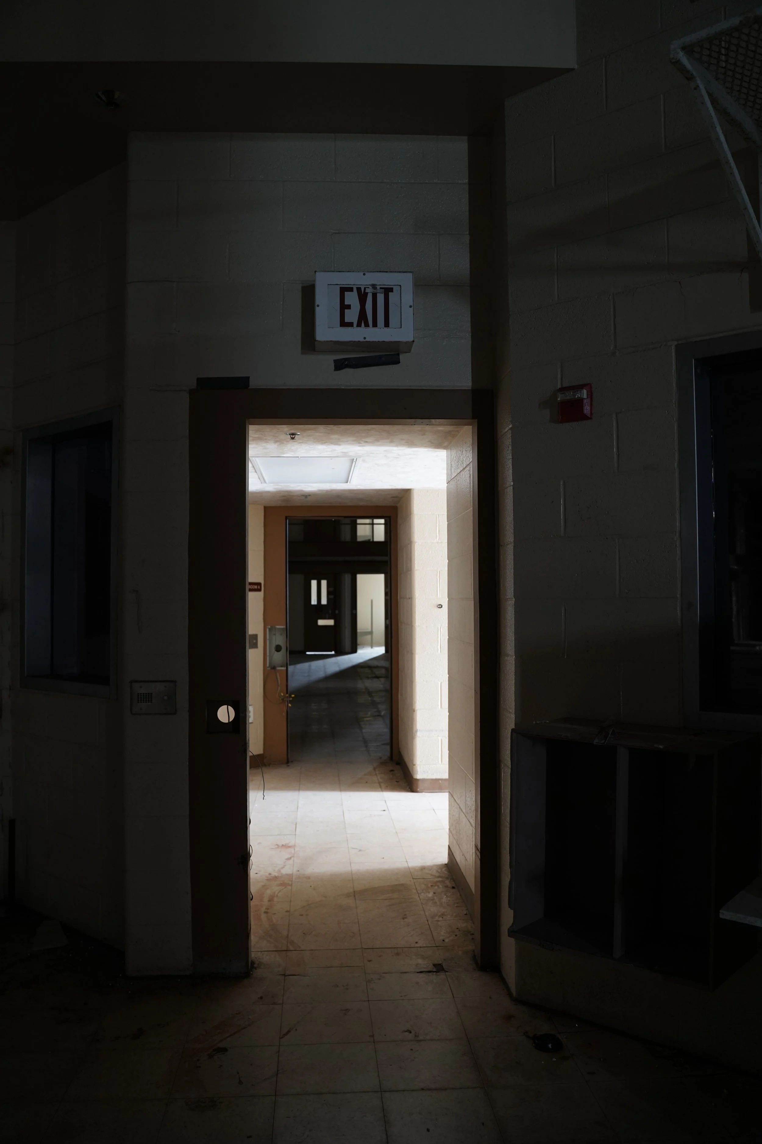 Dimly lit corridor inside a building with an illuminated exit sign overhead, leading to a brightly lit area with tiled floors and fluorescent ceiling lights.
