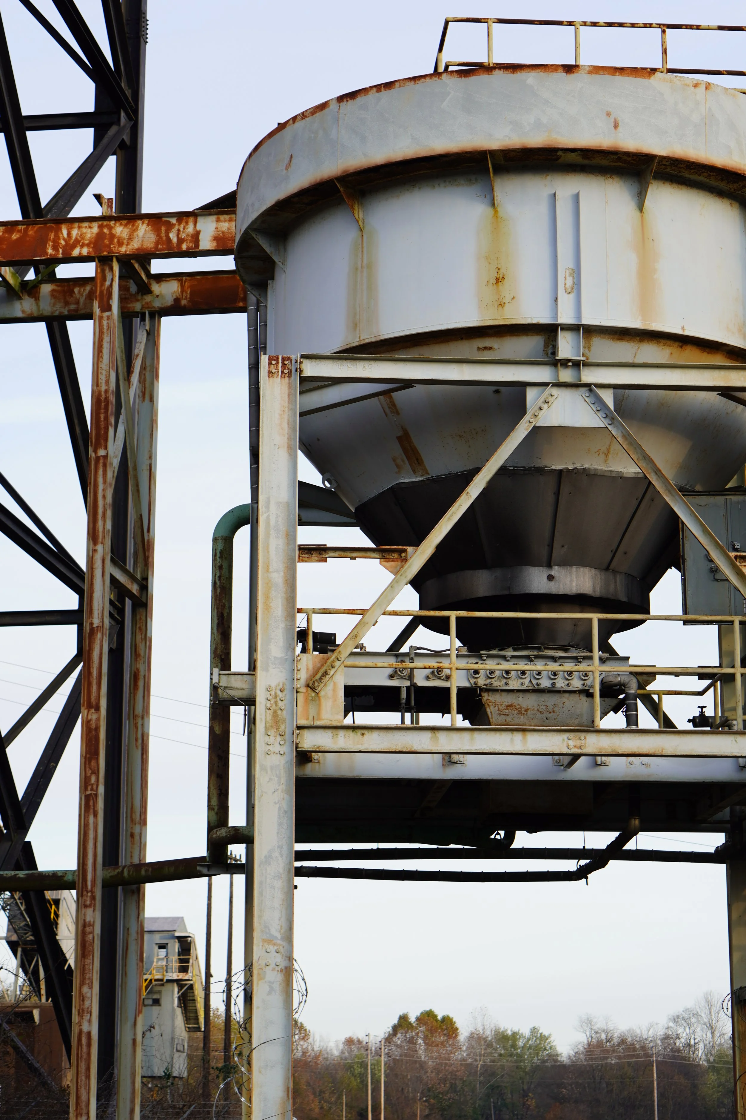 Rusty industrial structure with large cylindrical tanks, supporting framework, and barbed wire at the bottom, set against a background of trees and power lines.