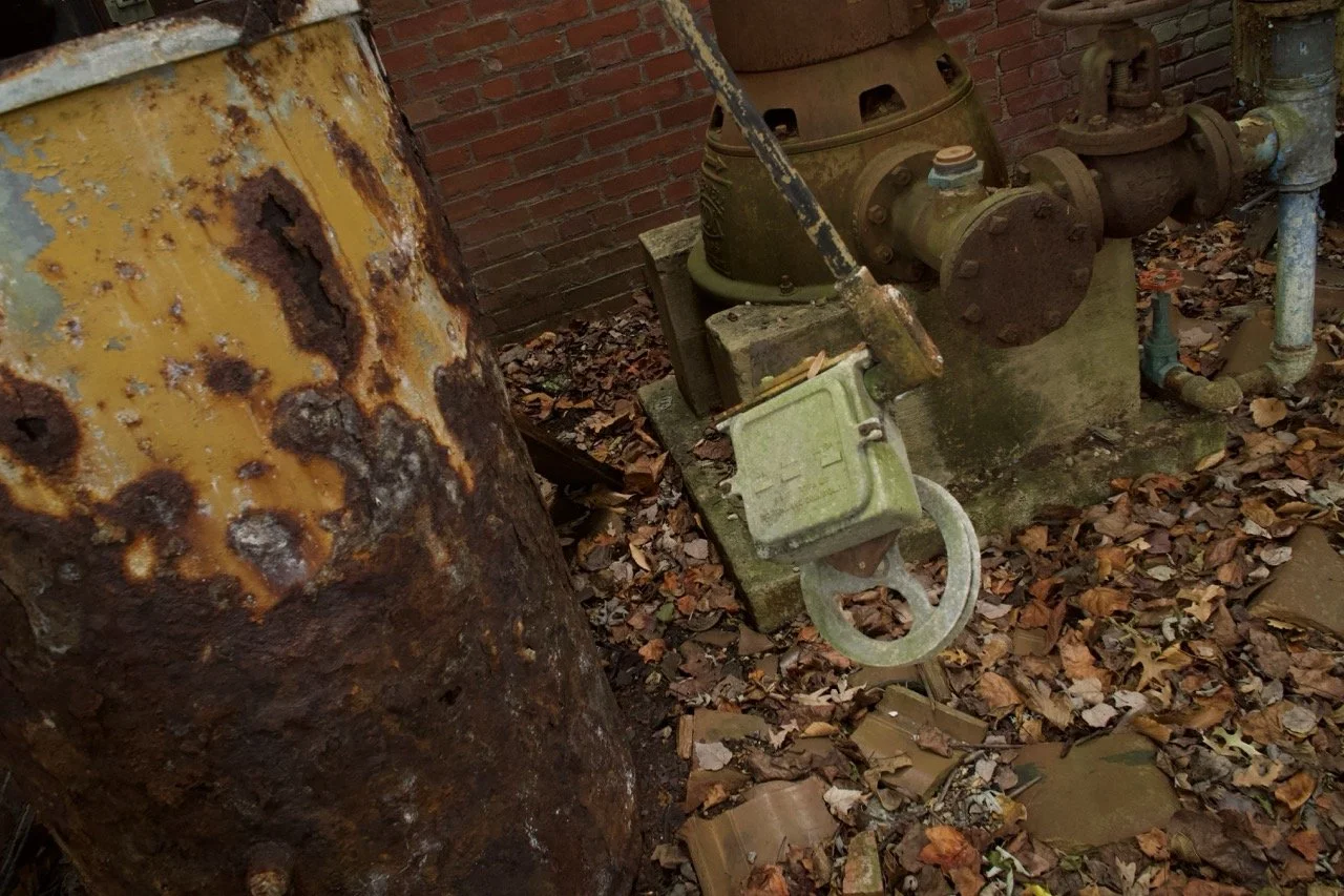Rusty, old industrial pipe and valve against a brick wall, surrounded by fallen dry leaves.