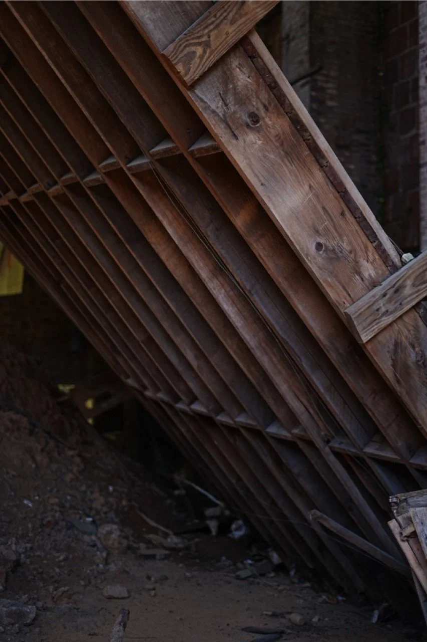 Vertical stack of wooden planks leaning against a brick wall, with soil and debris on the ground.