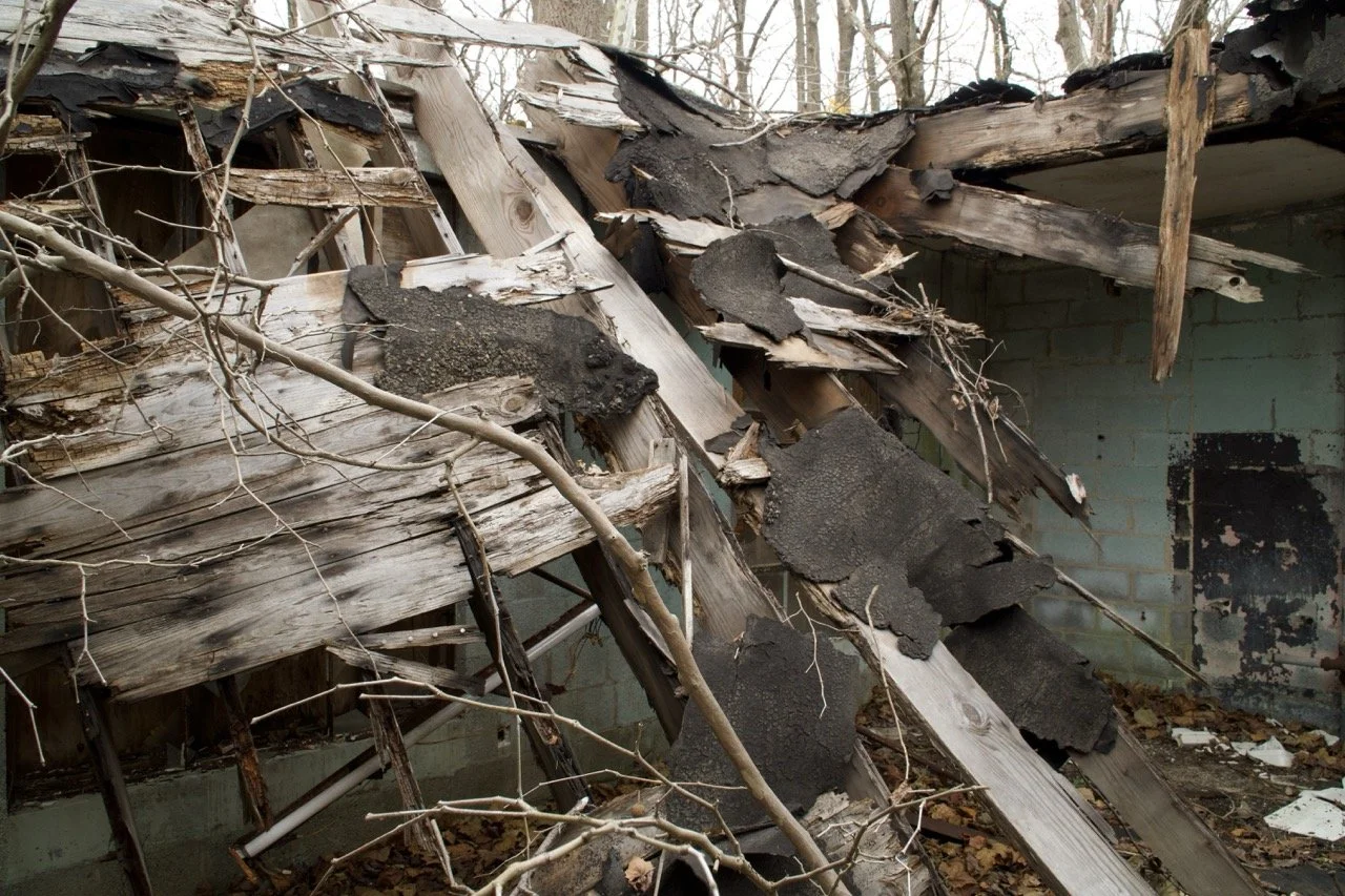 The image shows the collapsed roof of an old building with broken wooden beams, tar paper, and debris scattered around. The wall in the background is painted light green with peeling paint.
