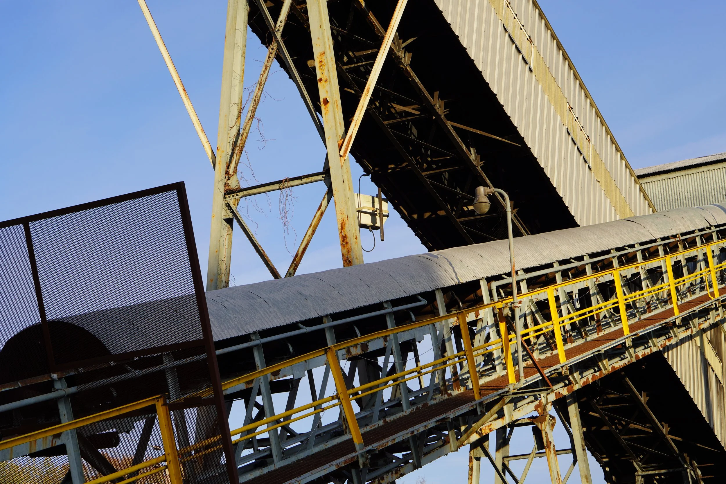 Close-up of an industrial structure with rusted metal beams, a covered conveyor system, and yellow safety railings against a blue sky.
