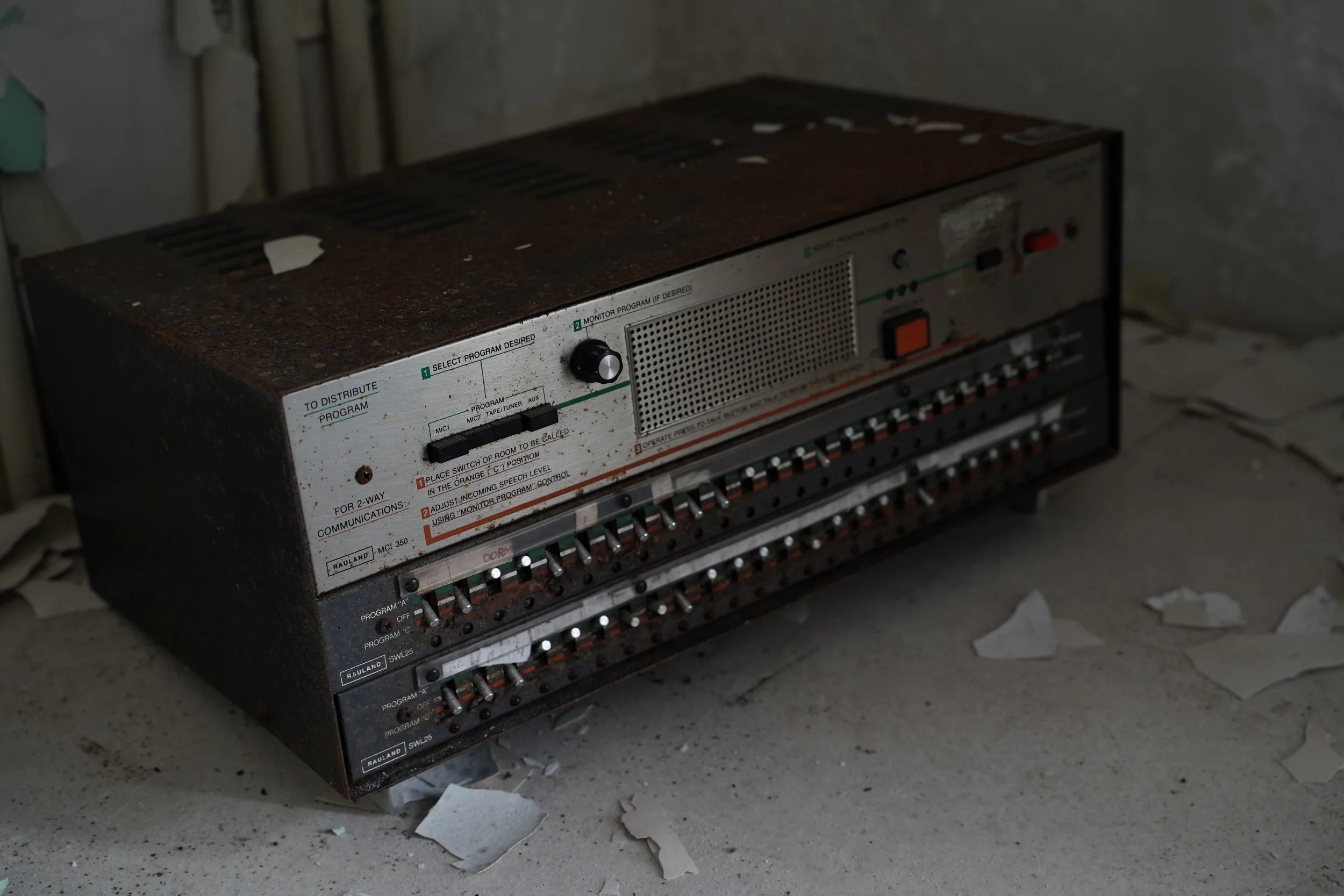 An old, rusty electronic device with numerous switches and labels, sitting on a concrete floor surrounded by scattered paper and debris.