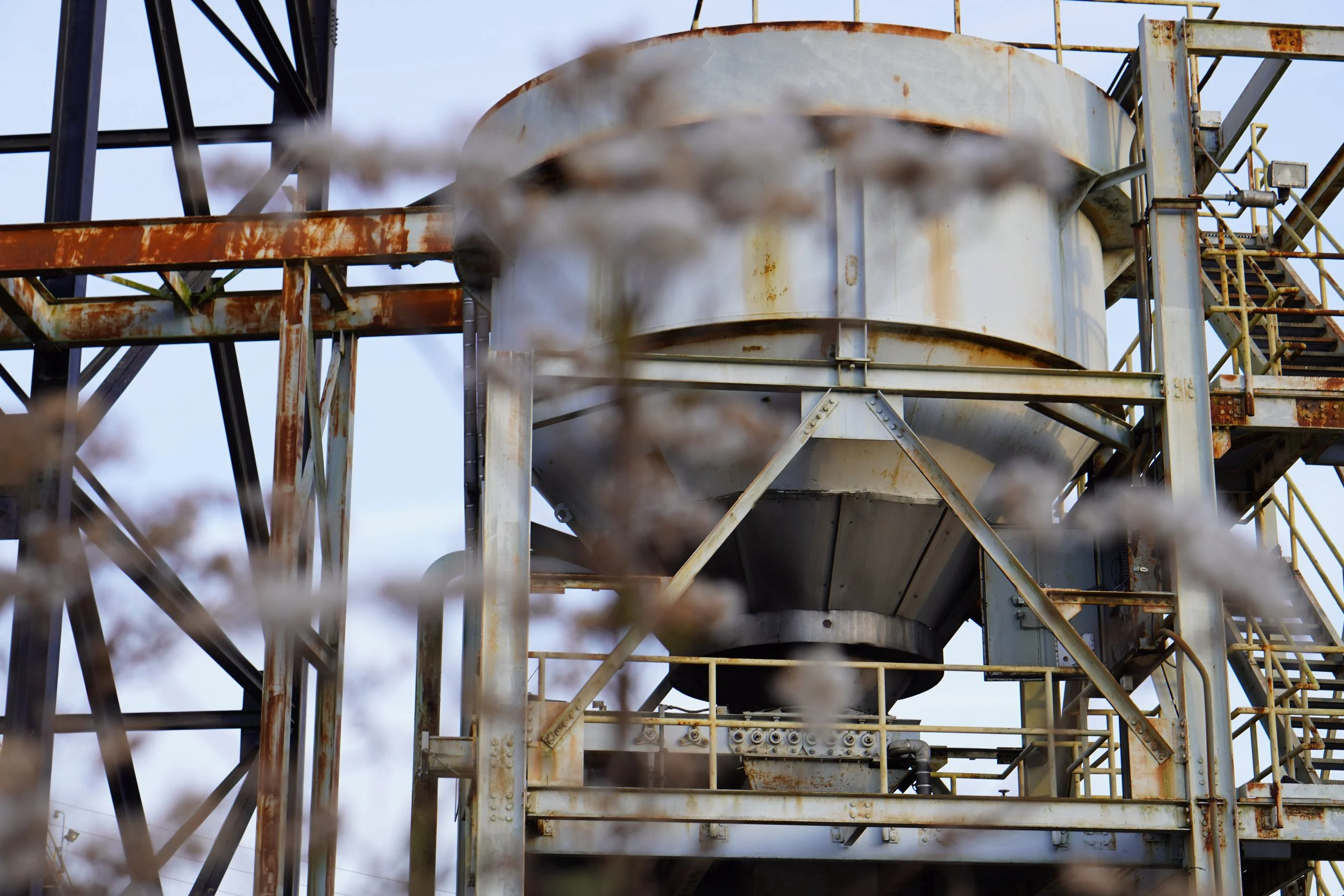 Rusty industrial metal structure with stairs, platforms, and pipes, with brown rust stains, against a cloudy sky background.