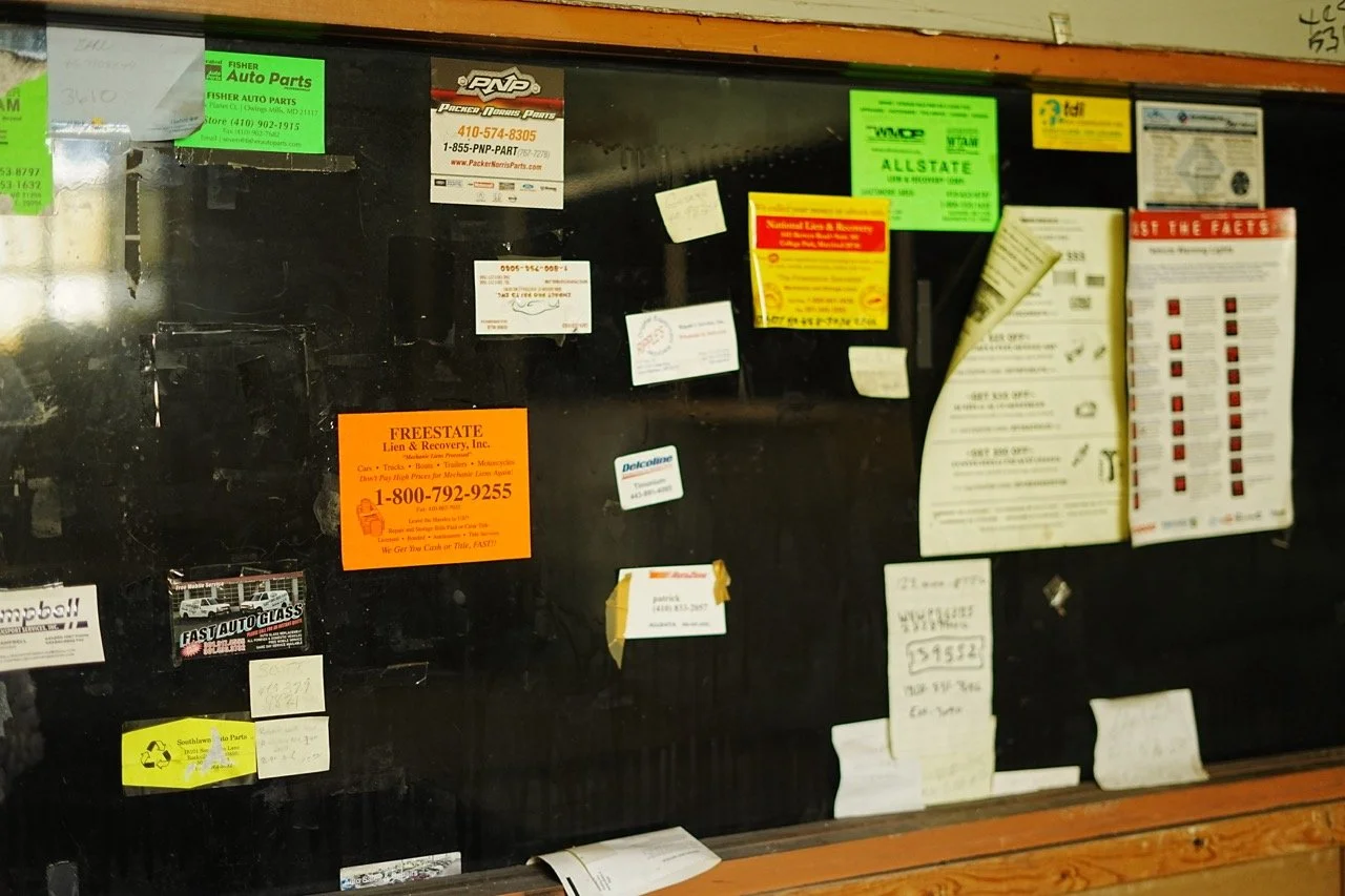 A bulletin board with various ads, business cards, flyers, and notices pinned to a black surface behind glass, with a wooden frame at the top and bottom.