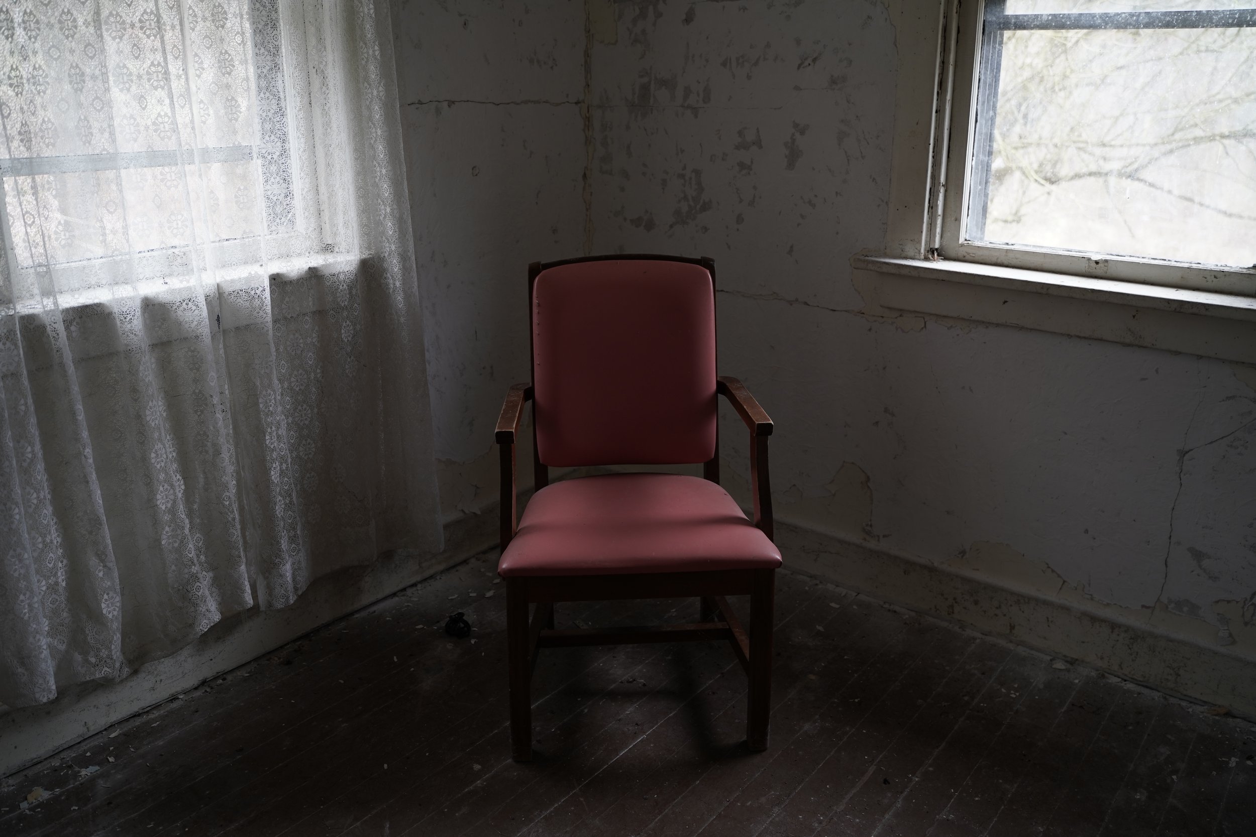 An old pink armchair with wooden arms and legs positioned in a corner of a decrepit room with peeling paint on the walls, a window with broken or dirty glass, and a lace curtain partially covering the window, with a dark wooden floor.