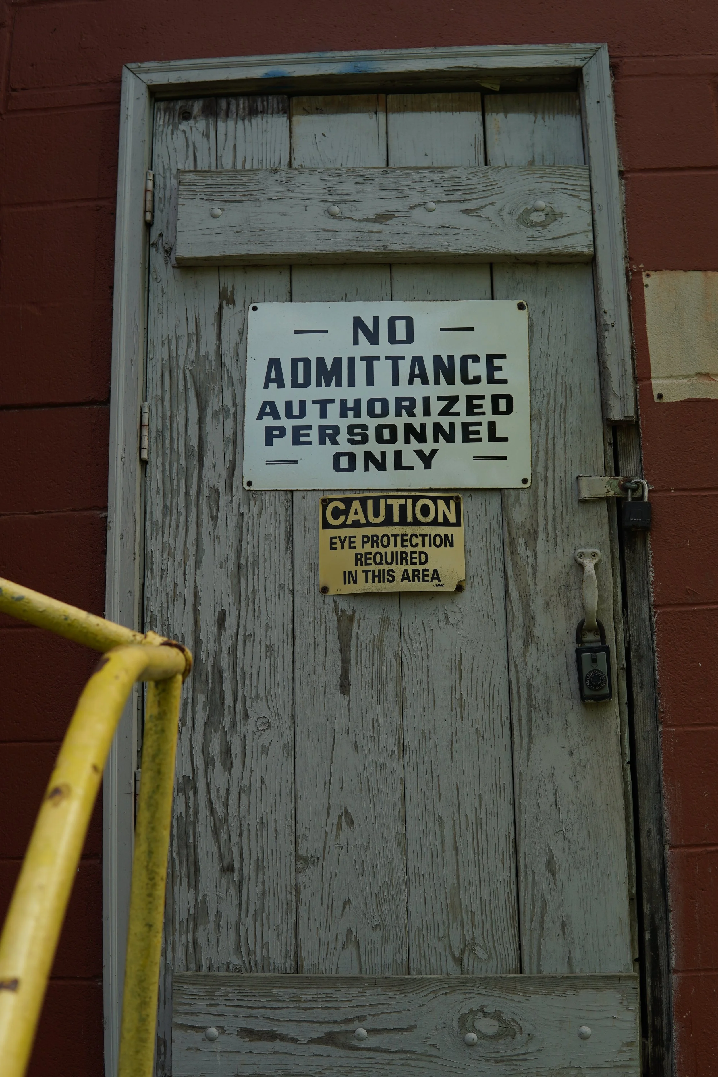 A weathered wooden door with a sign that reads 'No Admittance Authorized Personnel Only' and a smaller sign cautioning eye protection required. The door is set in a brick wall, with a yellow safety railing partially visible in the foreground.
