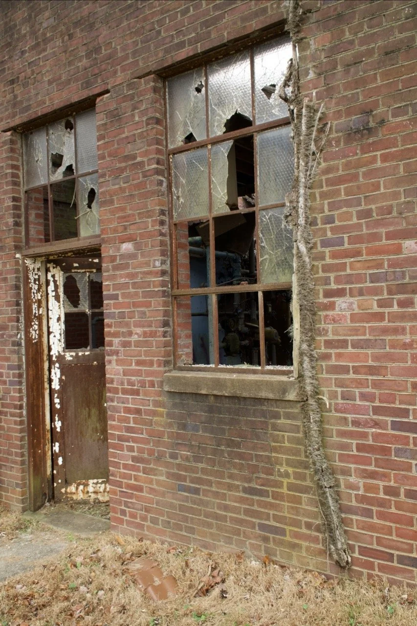 Broken glass windows on an old brick building, with a damaged door and a large crack running down the brick wall.