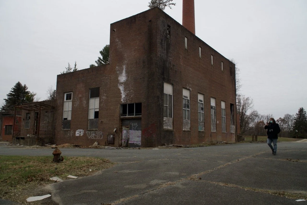An old, abandoned brick building with boarded-up and broken windows, graffiti on the walls, and a tall brick chimney, situated in a desolate area with a person wearing a mask walking nearby.
